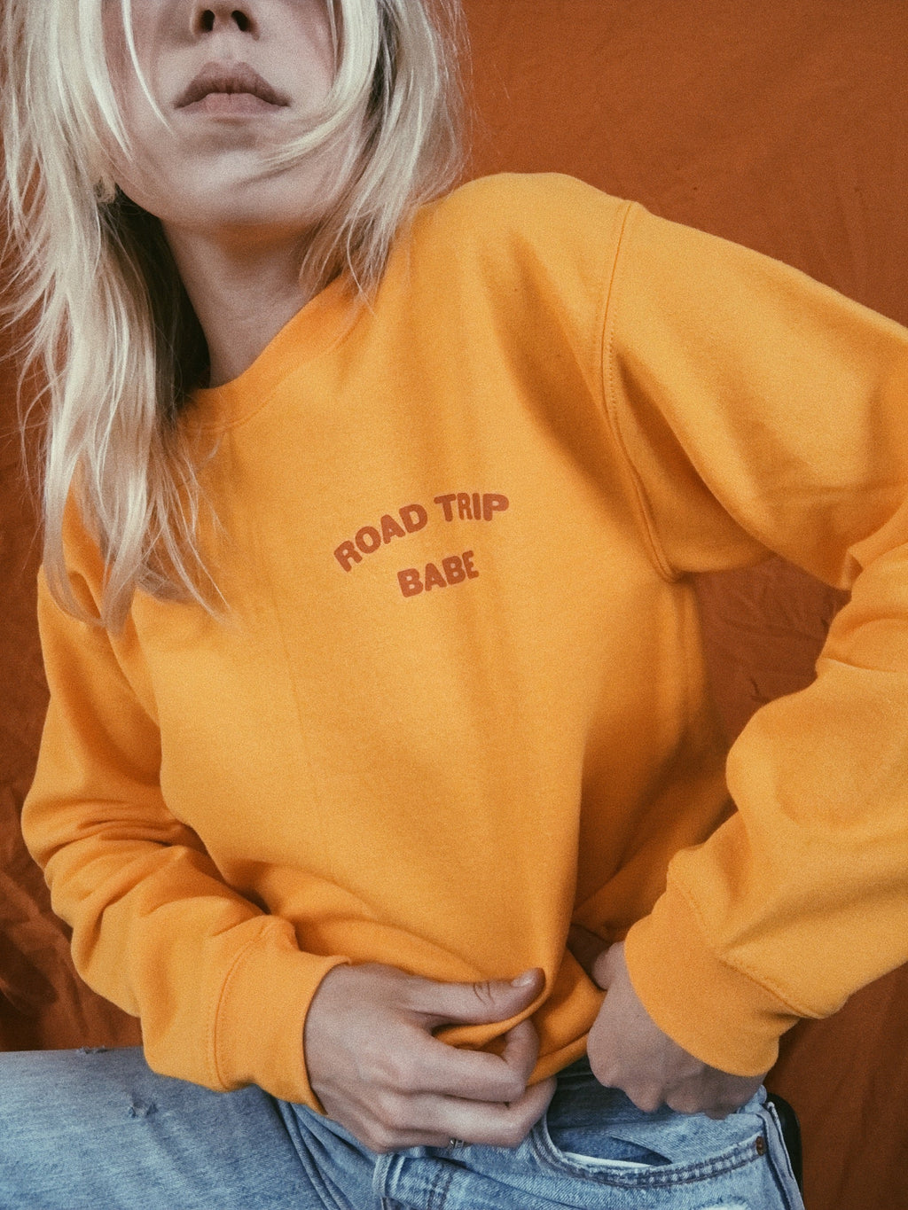 Yellow crewneck sweatshirt with text 'ROAD TRIP BABE' on corner of chest being worn against a vintage orange backdrop