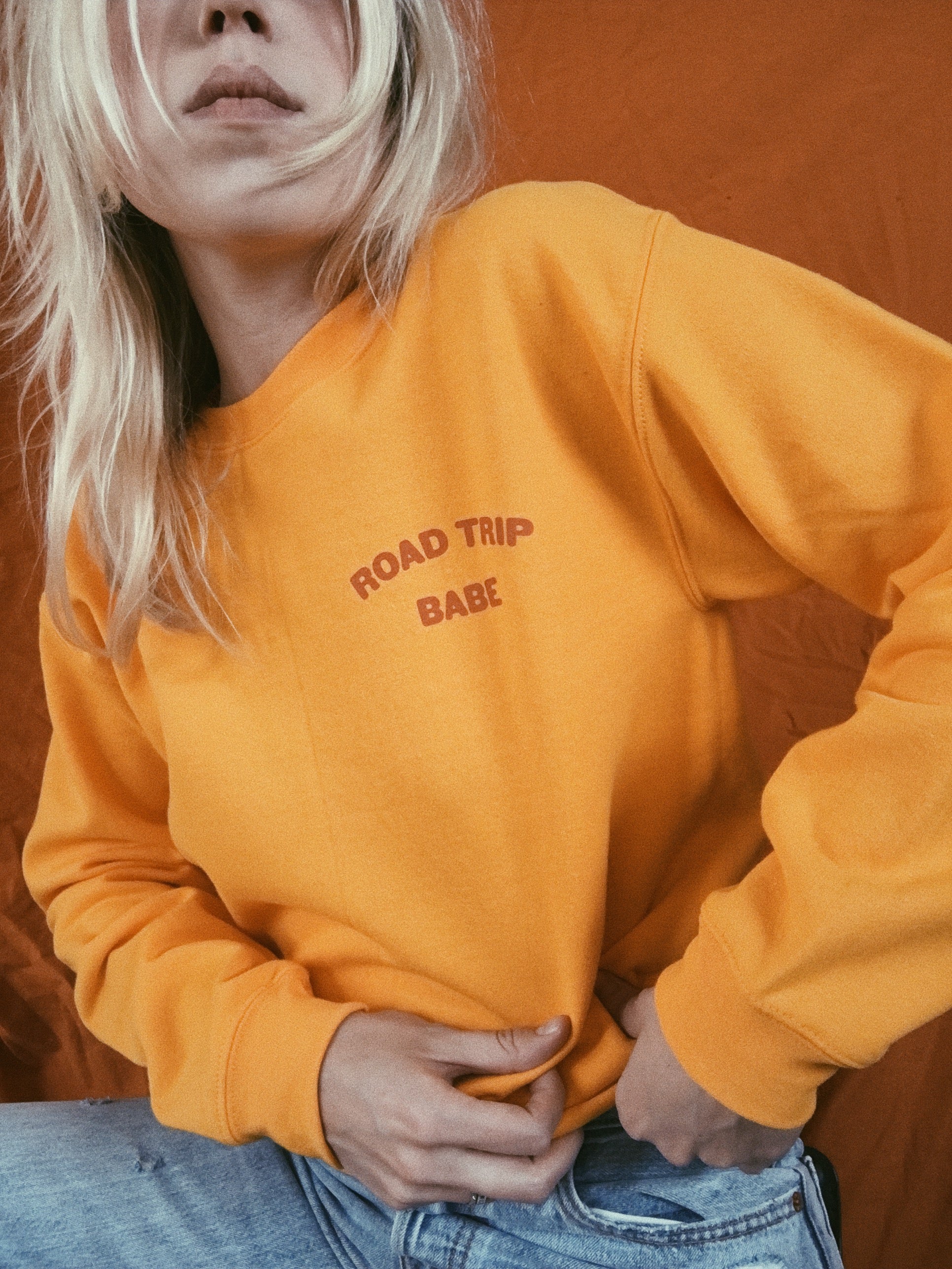 Yellow crewneck sweatshirt with text 'ROAD TRIP BABE' on corner of chest being worn against a vintage orange backdrop