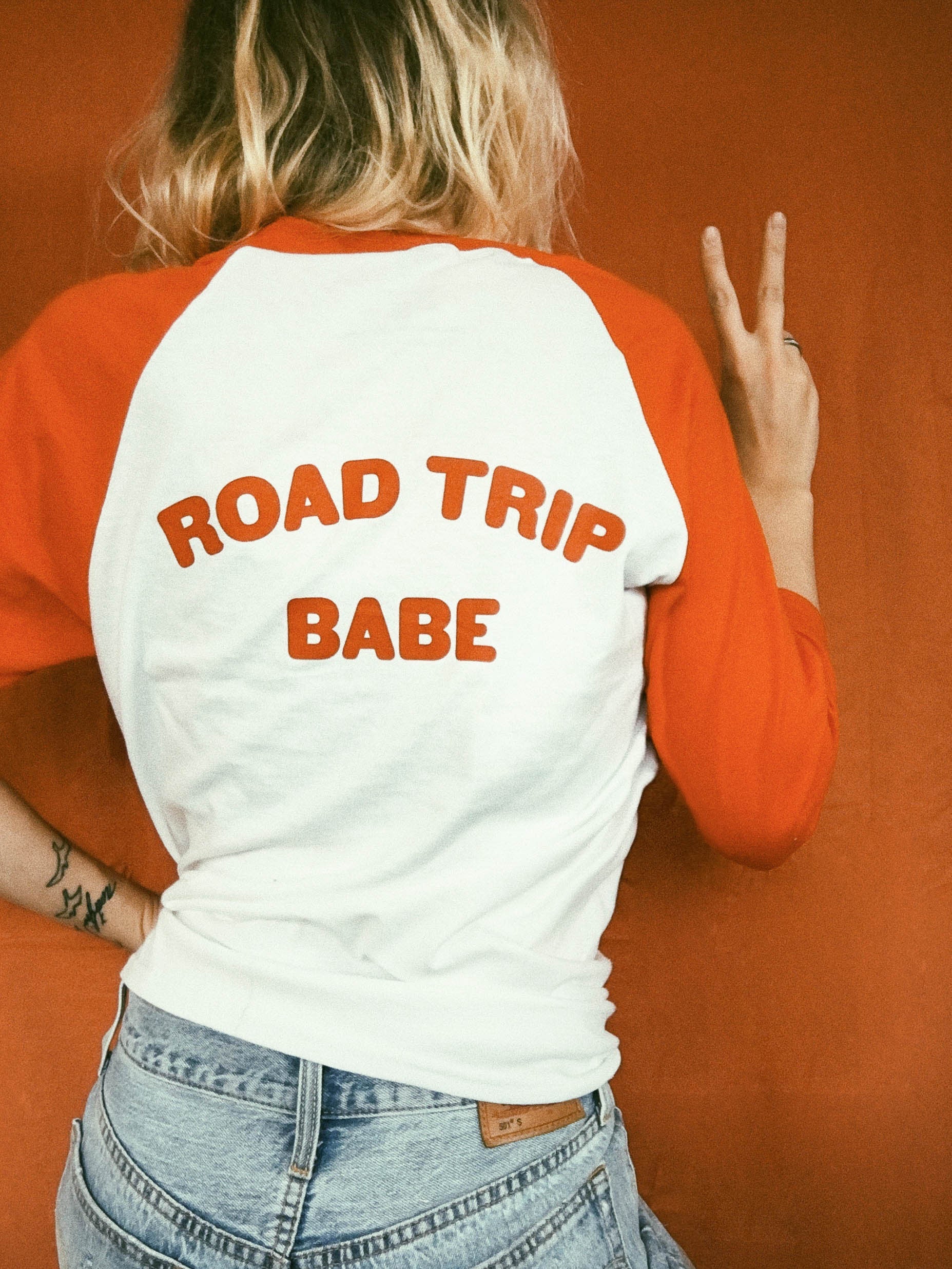 Red and white raglan tee with text 'ROAD TRIP BABE' in red ink screen printed across the back being worn against a vintage orange backdrop