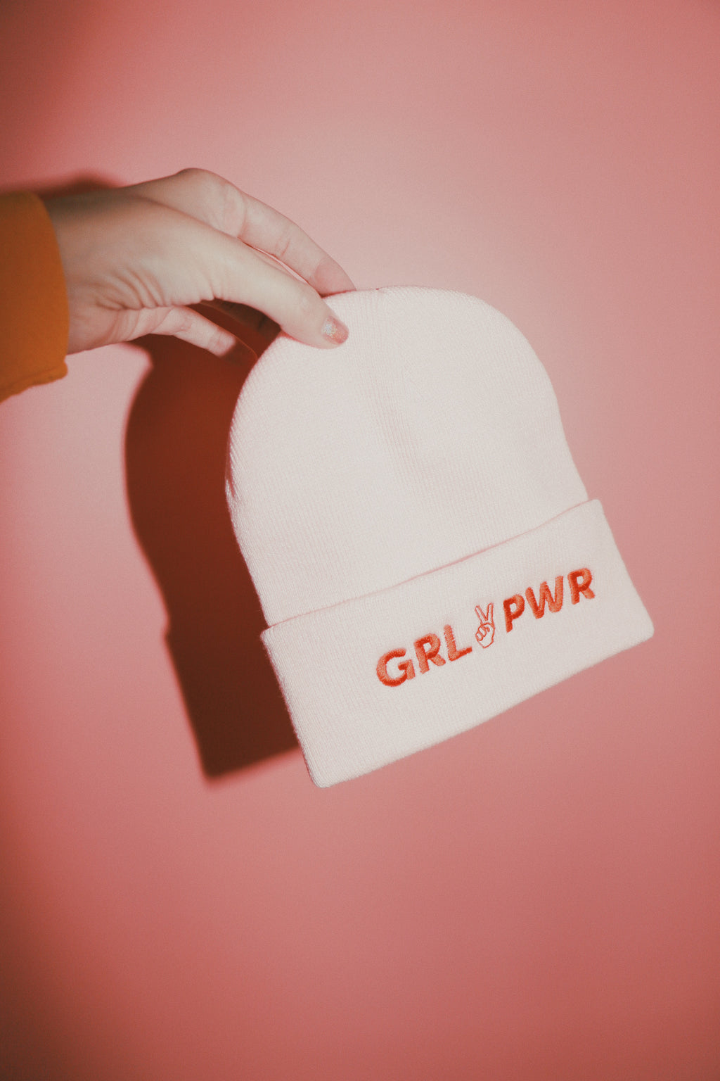 light pink beanie with text 'GRL PWR' in red thread embroidered across band of the beanie being held against a pink backdrop
