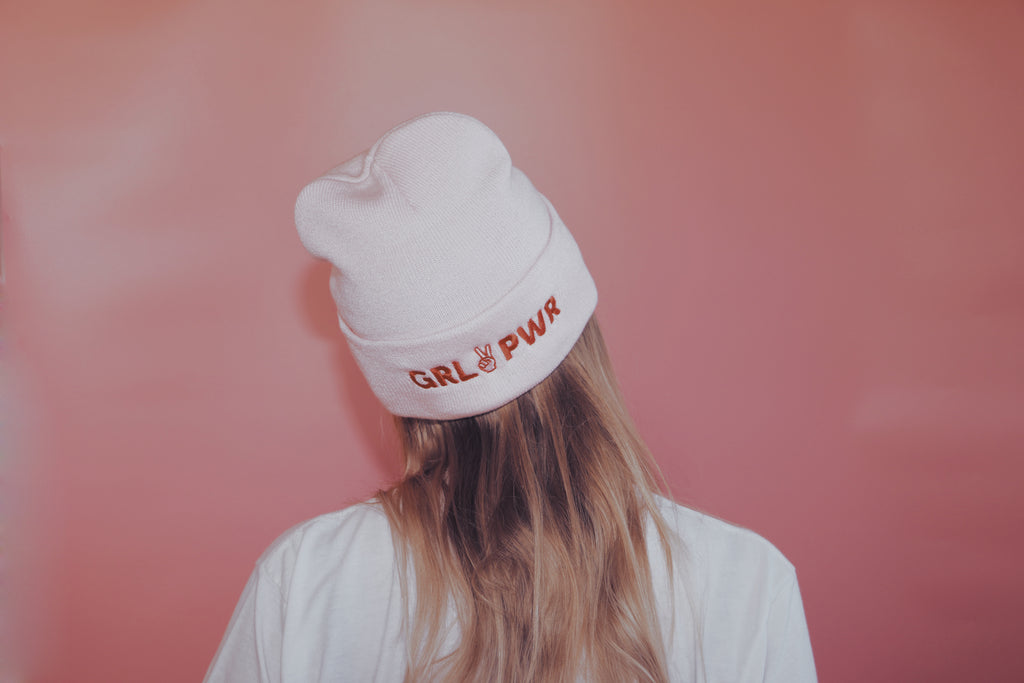 light pink beanie with text 'GRL PWR' in red thread embroidered across band of the beanie being worn in front on a pink backdrop