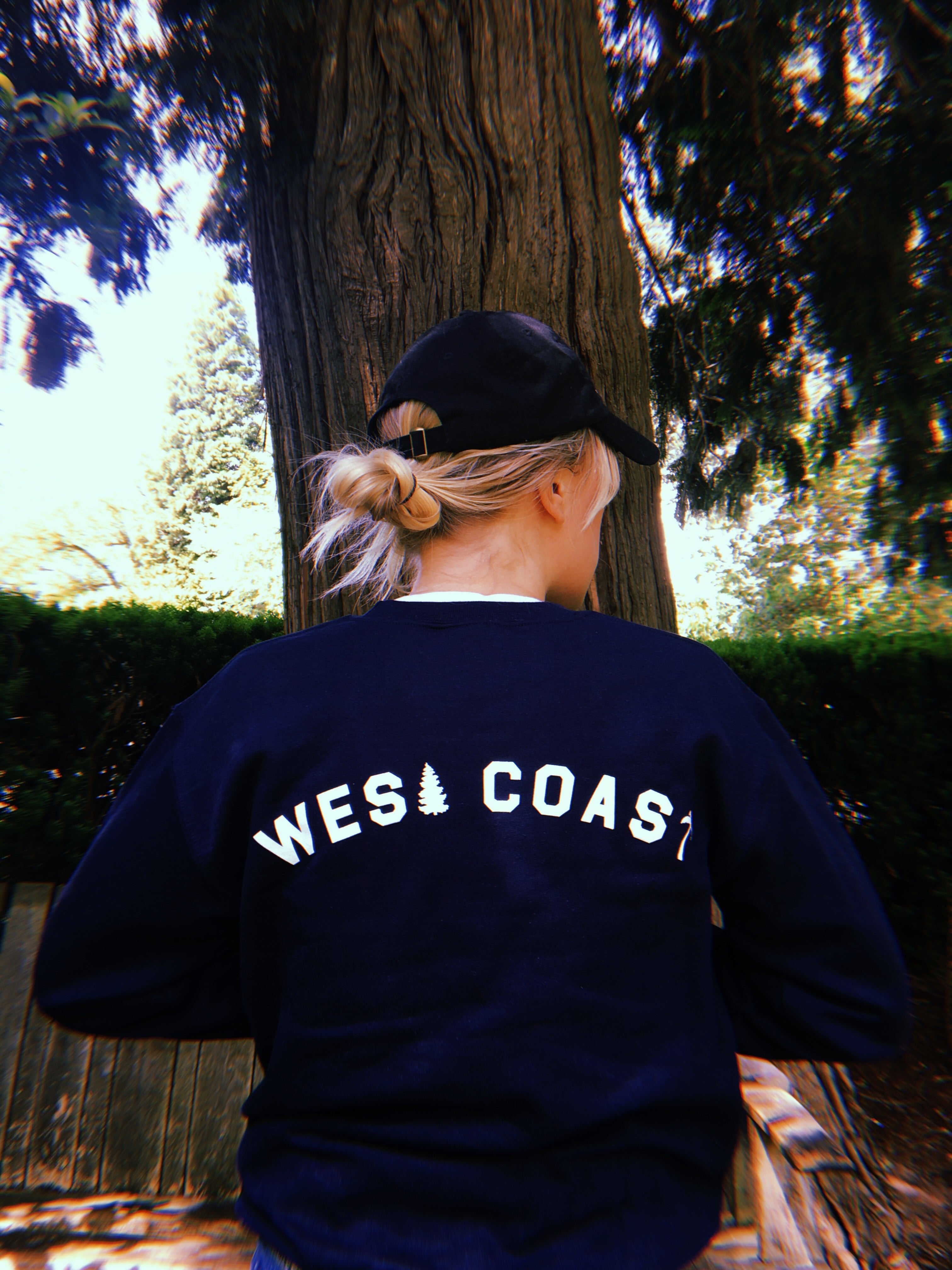 navy blue crewneck with text 'WEST COAST' text across the back being worn in forest area