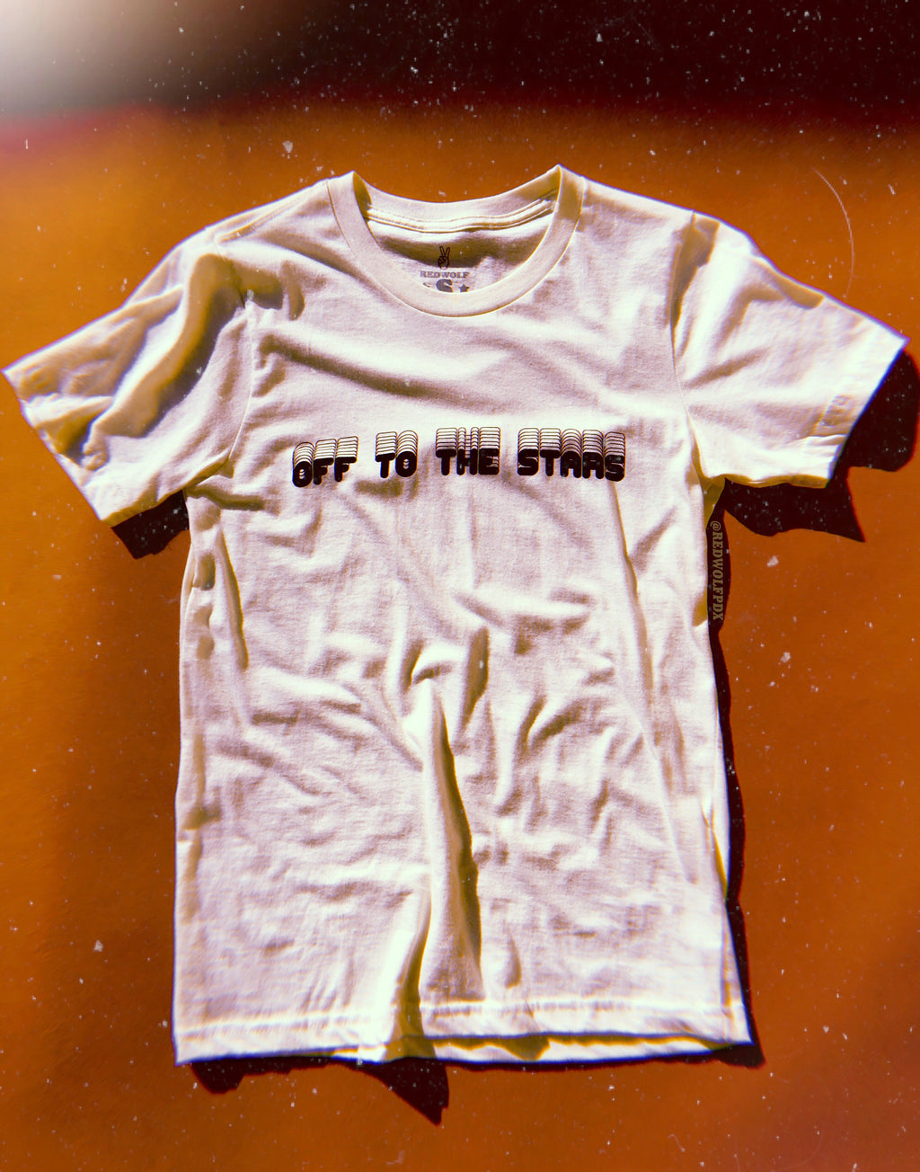 white tee with  text 'OFF TO THE STARS' in vintage inspired font screen printed in black ink across chest against a brown background
