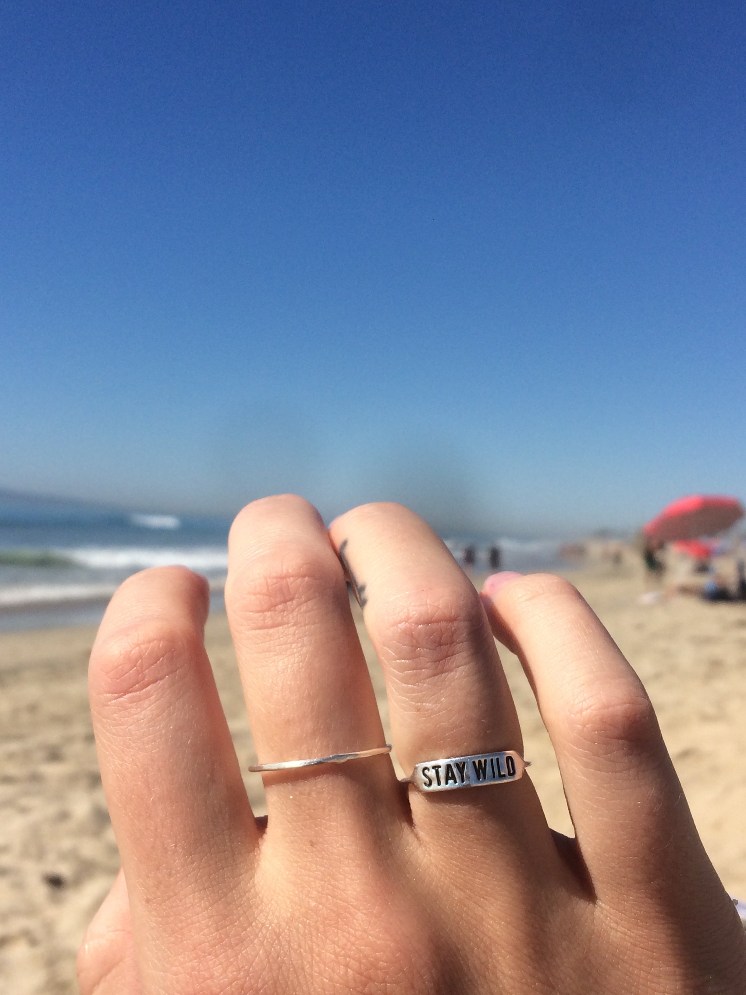 sterling silver ring with text 'STAY WILD' engraved on it being worn on hand with beach background
