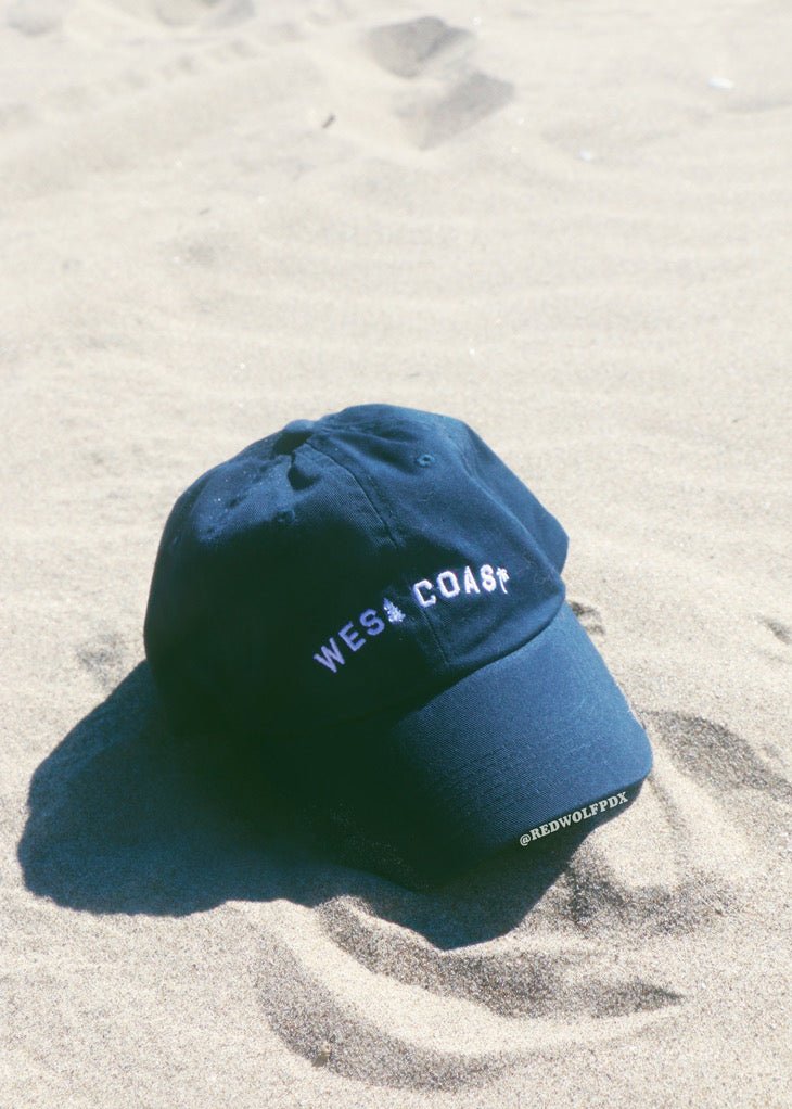 Navy blue baseball cap with text 'WEST COAST' embroidered across rested on sandy beach