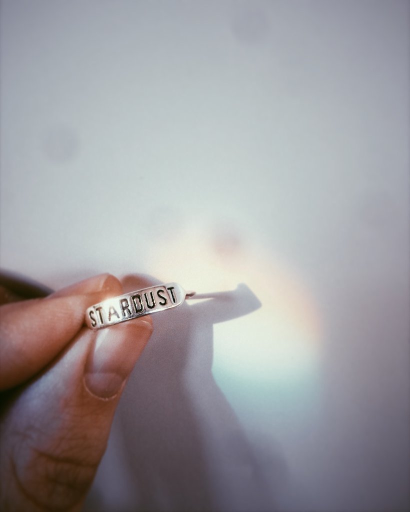 a sterling silver ring with text 'STARDUST' engraved on it being held up by a hand to a white background with a rainbow prism