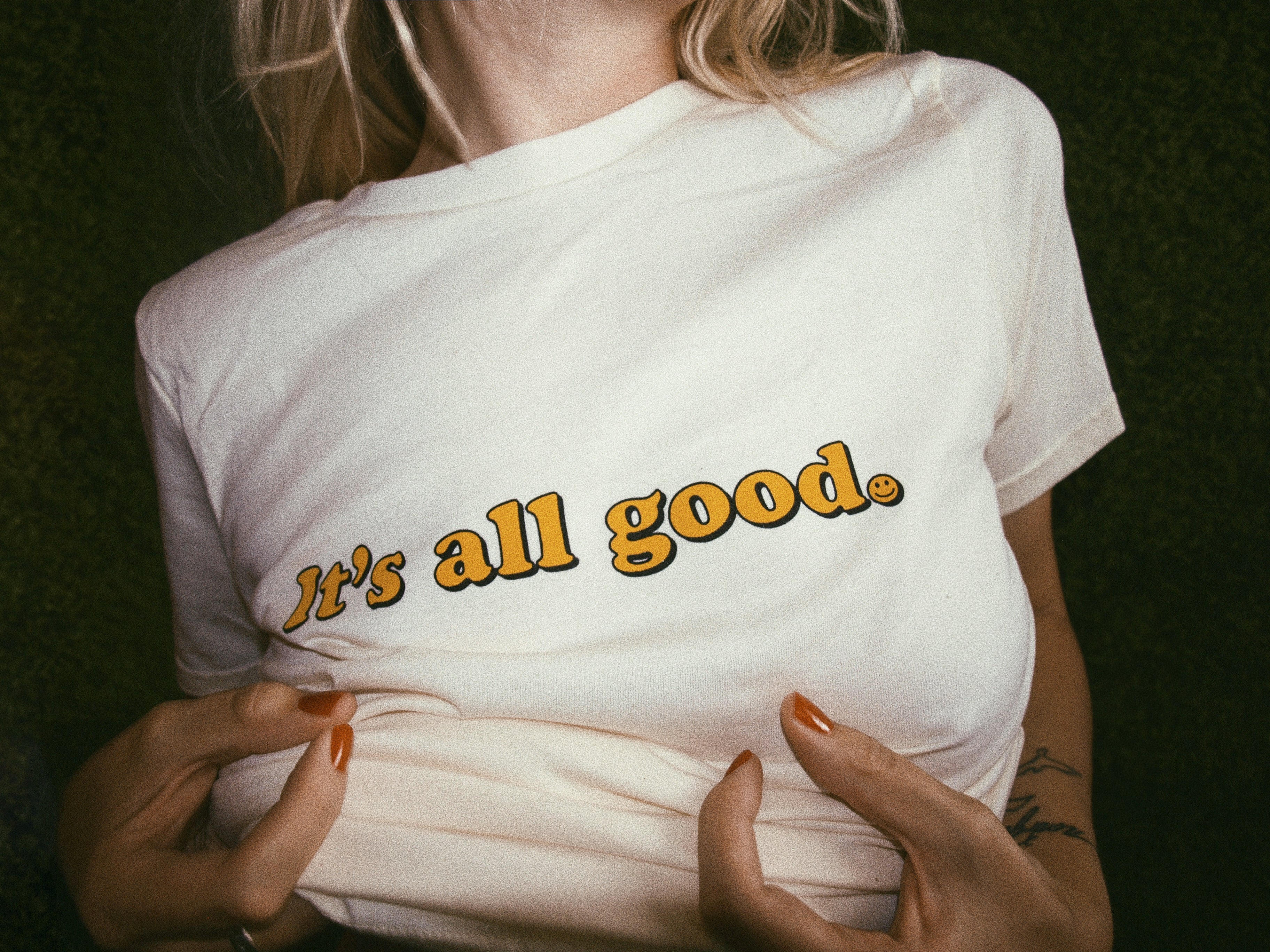 Beige white graphic tee with text 'It's all good' with smiley face in yellow ink screen printed across chest being worn with green backdrop