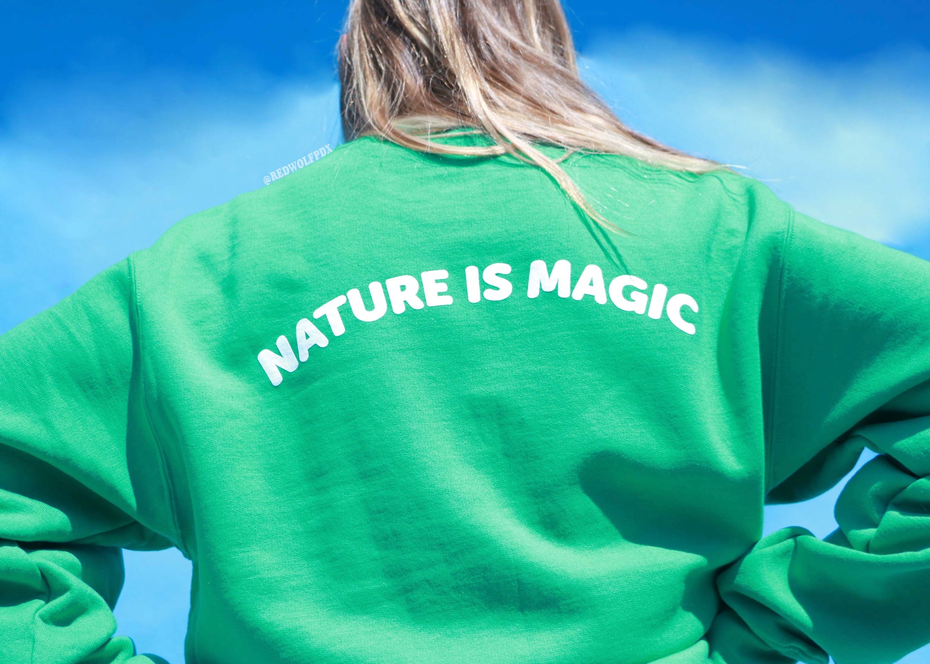 Green crewneck sweatshirt with the text 'NATURE IS MAGIC' printed across the back being worn against a blue backdrop