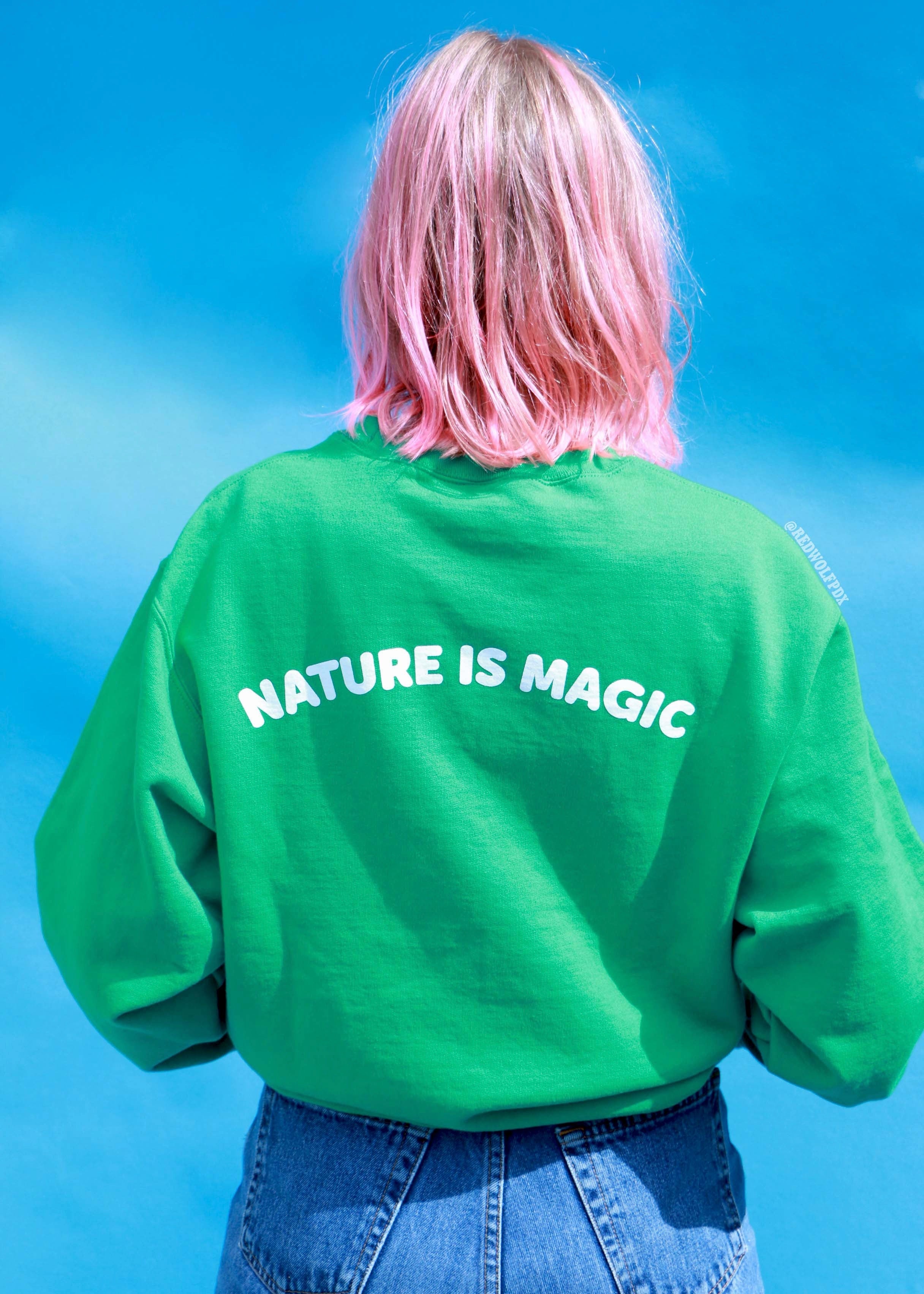 Green crewneck sweatshirt with the text 'NATURE IS MAGIC' printed across the back being worn against a blue backdrop