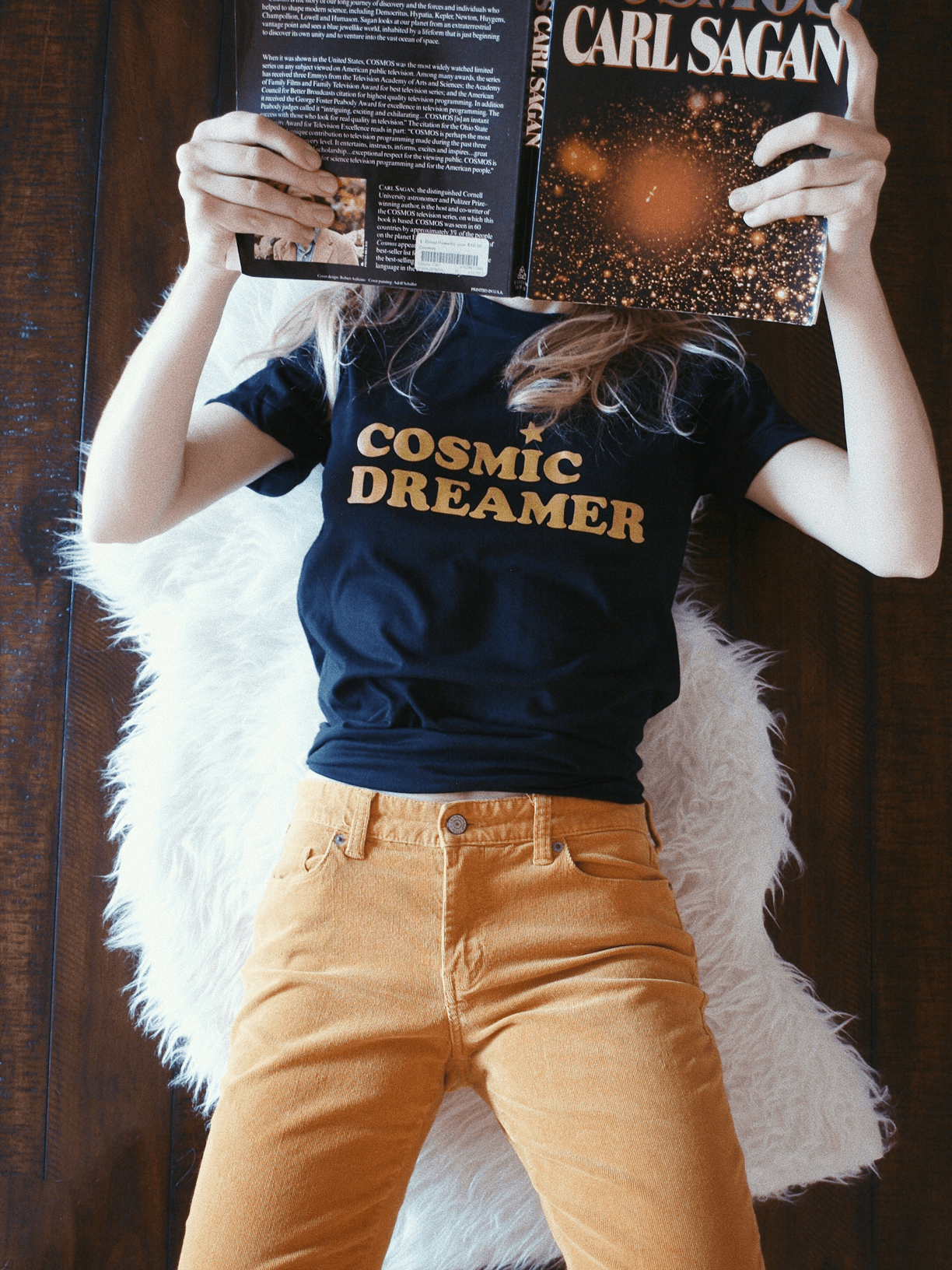 black graphic tee with text 'COSMIC DREAMER' in yellow ink screen printed across chest being worn while holding up a book on a wood floor 