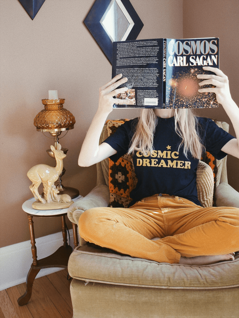 black graphic tee with text 'COSMIC DREAMER' in yellow ink across chest being worn in a green velvet chair holding up a book in corner of a room.