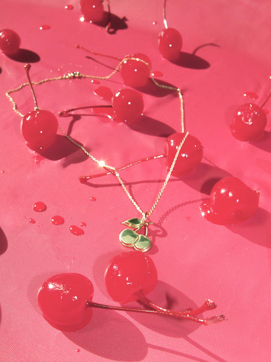A brass  necklace with a pendant shaped like a cherry, displayed on a pink backdrop  with red cherries and cherry syrup droplets.