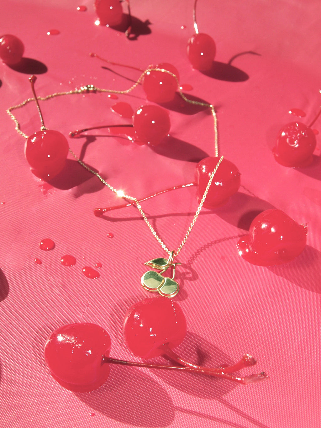 A brass  necklace with a pendant shaped like a cherry, displayed on a pink backdrop  with red cherries and cherry syrup droplets. 
