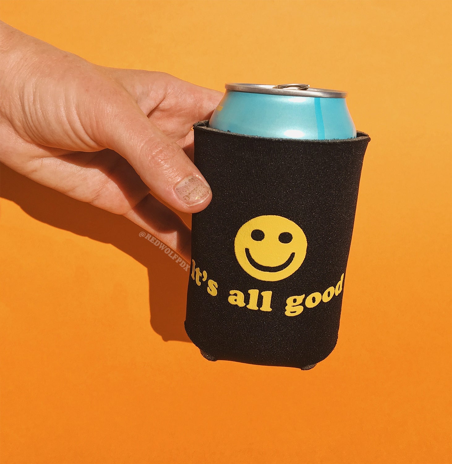 12 oz can beverage in a black neoprene drink sleeve with text 'It's all good' and smiley face graphic in yellow ink screen printed across it being held against an orange backdrop