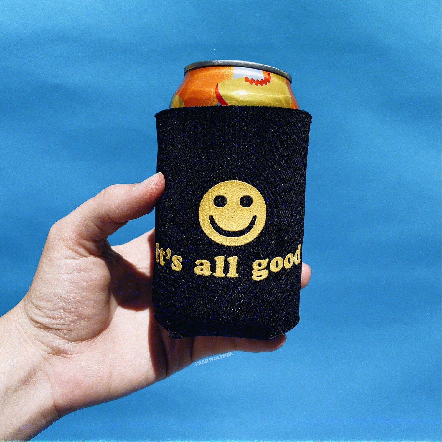 12 oz can in black neoprene drink sleeve with text 'It's all good' with smiley face in yellow ink screen printed on it being held against blue backdrop