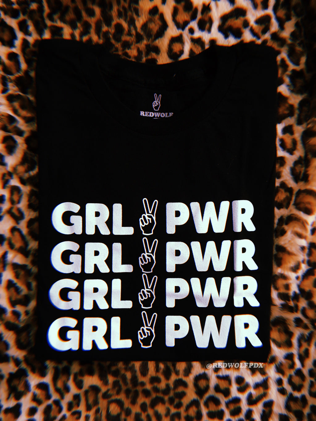 folded black tee with text 'GRL PWR' with peace sign graphic screen printed down the front in white ink on a leopard print background