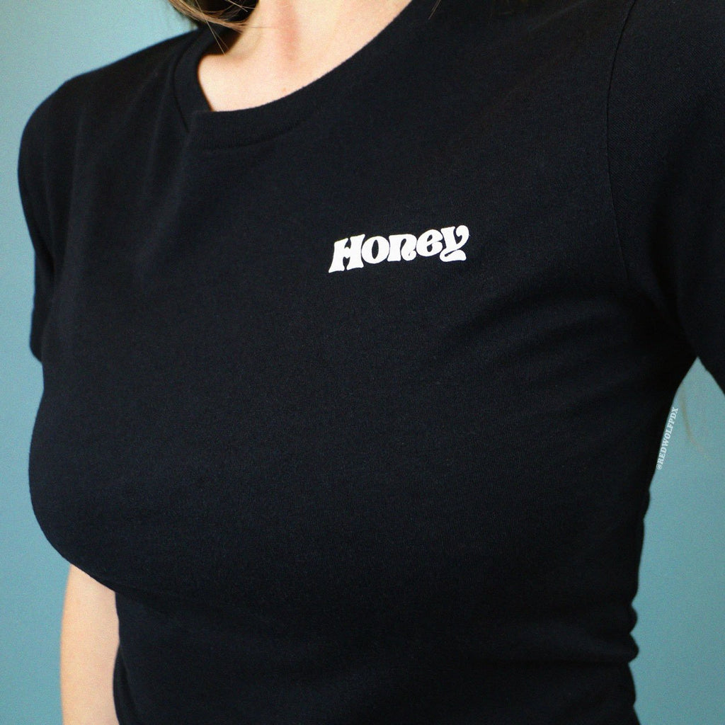 Black fitted graphic tee with the text 'Honey' on corner of chest being worn against blue backdrop