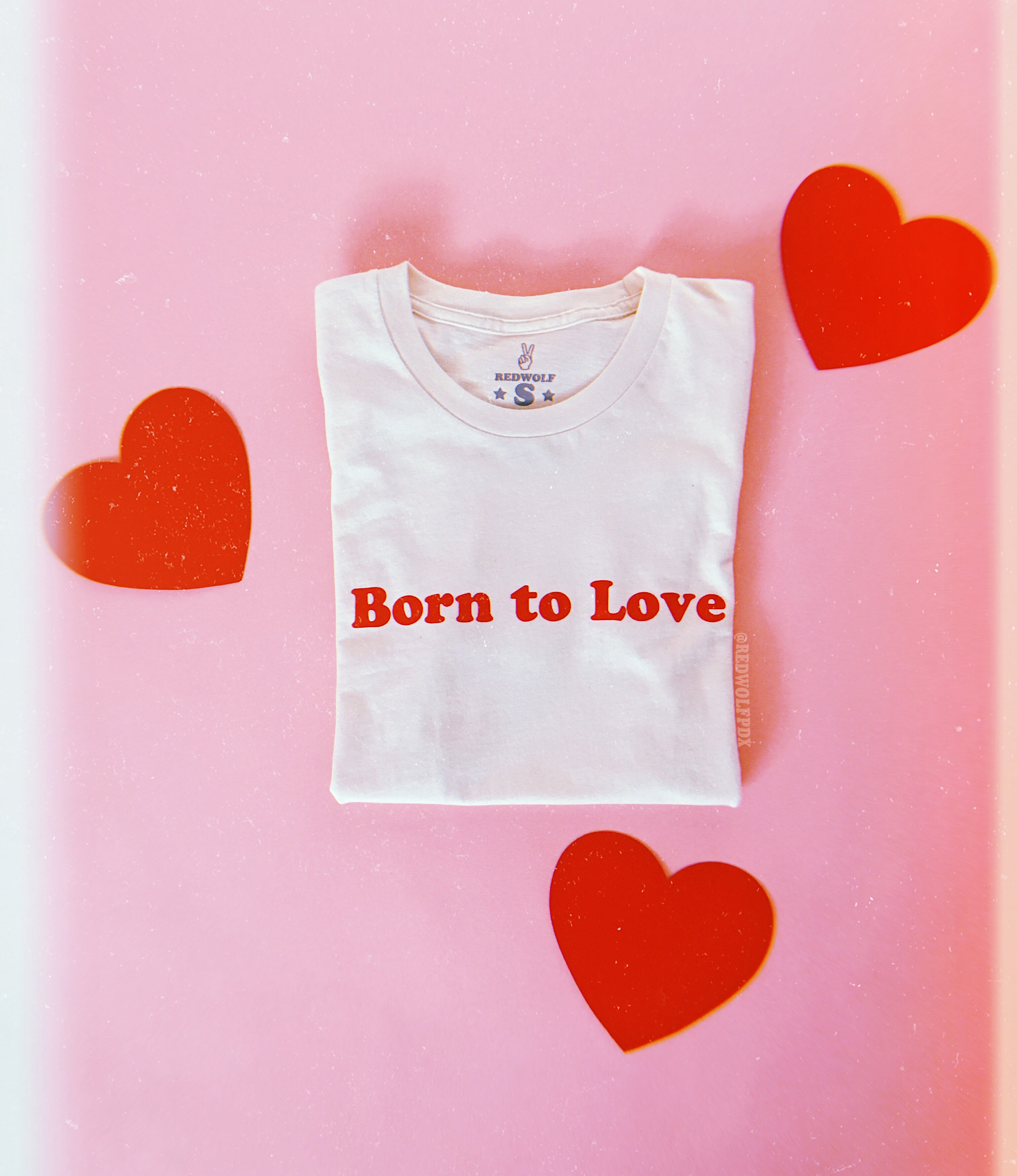 A white crew neck t-shirt with the red graphic 'Born to Love' printed across the front, displayed on a pink background with red hearts