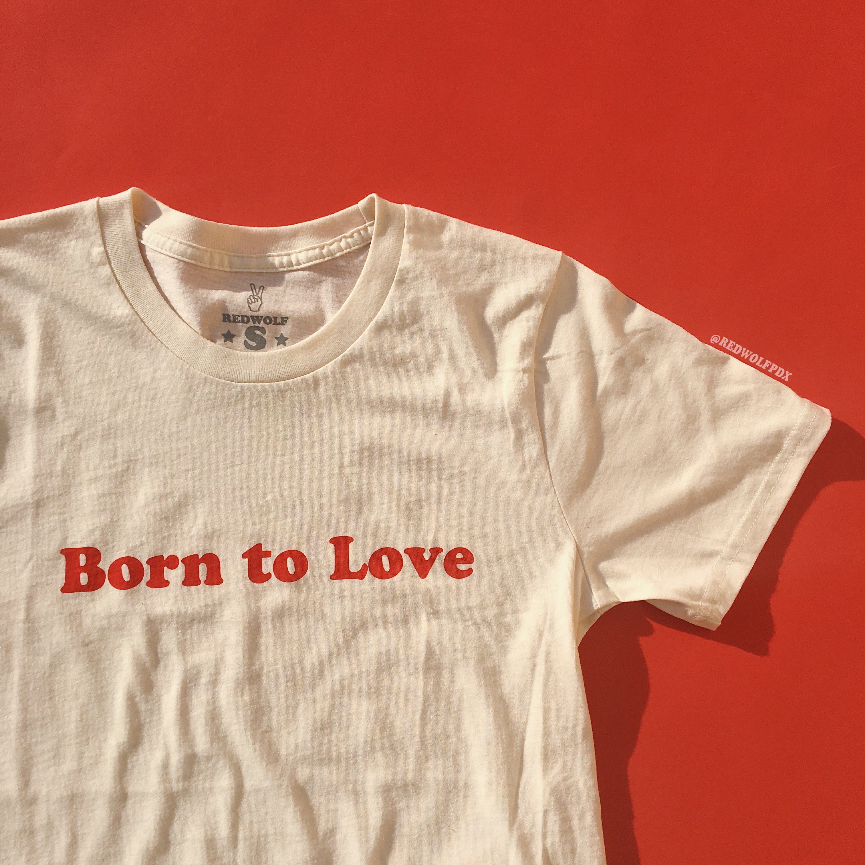 
A white crew neck t-shirt with the red graphic 'Born to Love' printed across the front, displayed on a red background.