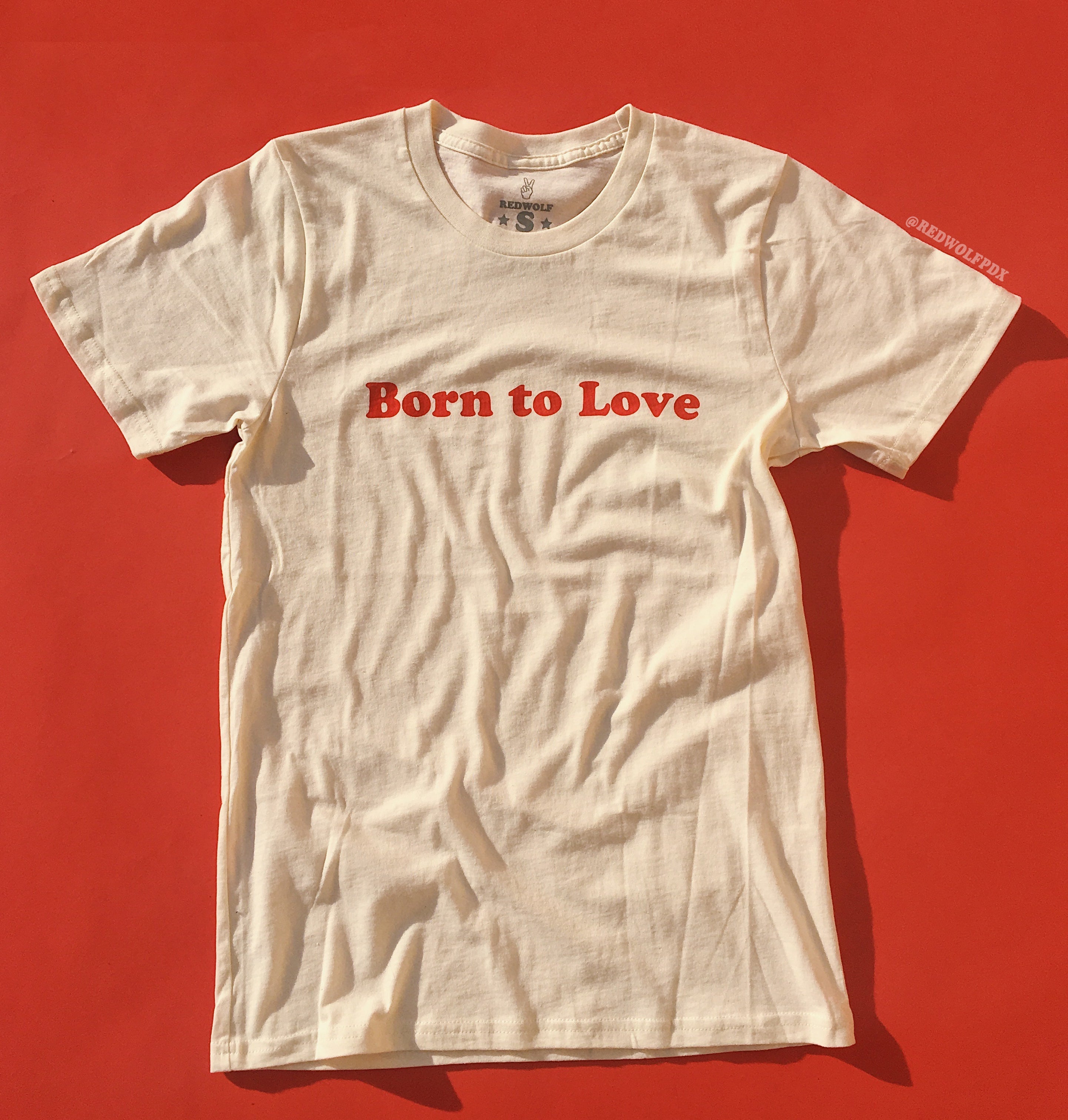 


A white crew neck t-shirt with the red graphic 'Born to Love' printed across the front, displayed on a red background.