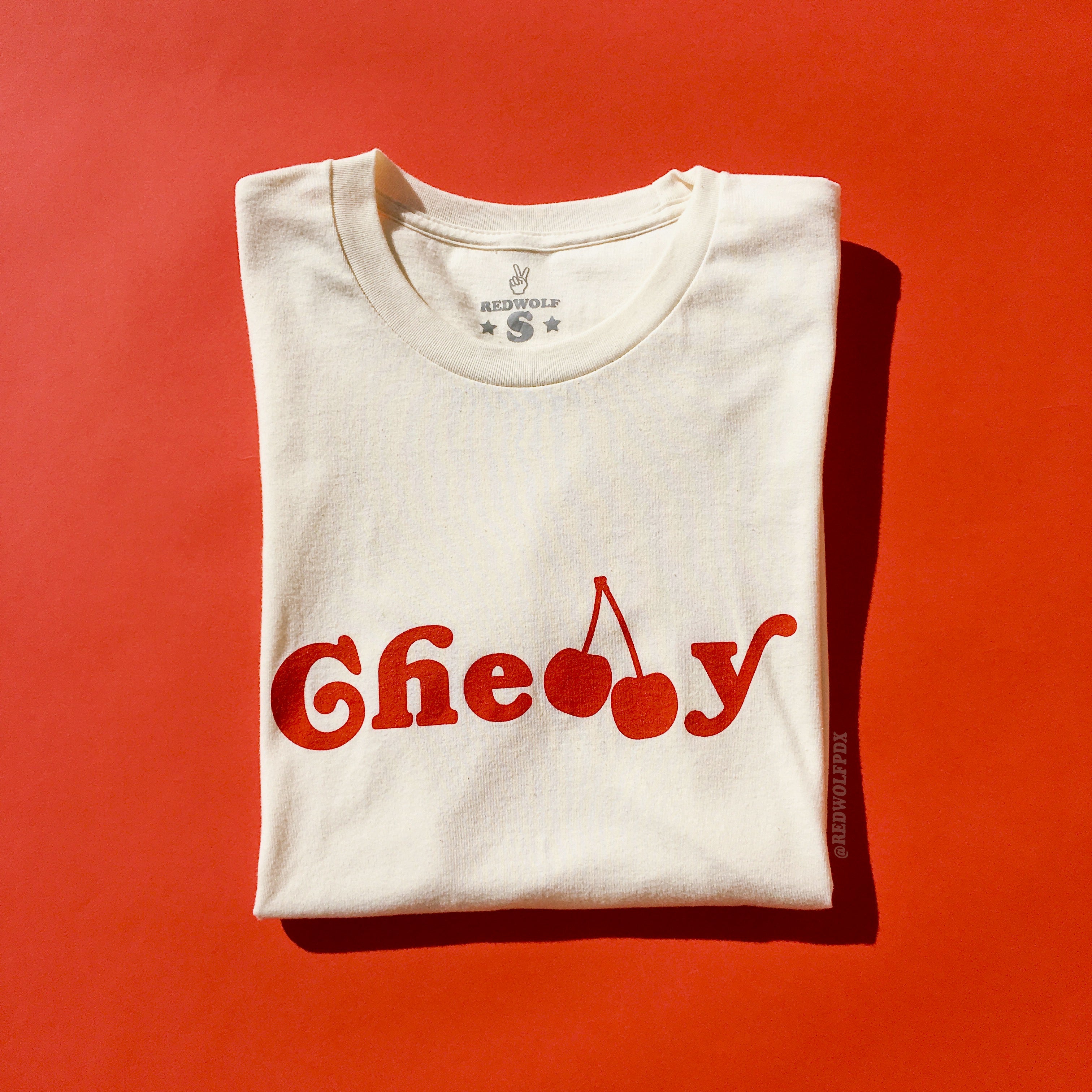 Folded beige white graphic tee with text 'Cherry' screen printed across in red ink on a vintage red backdrop