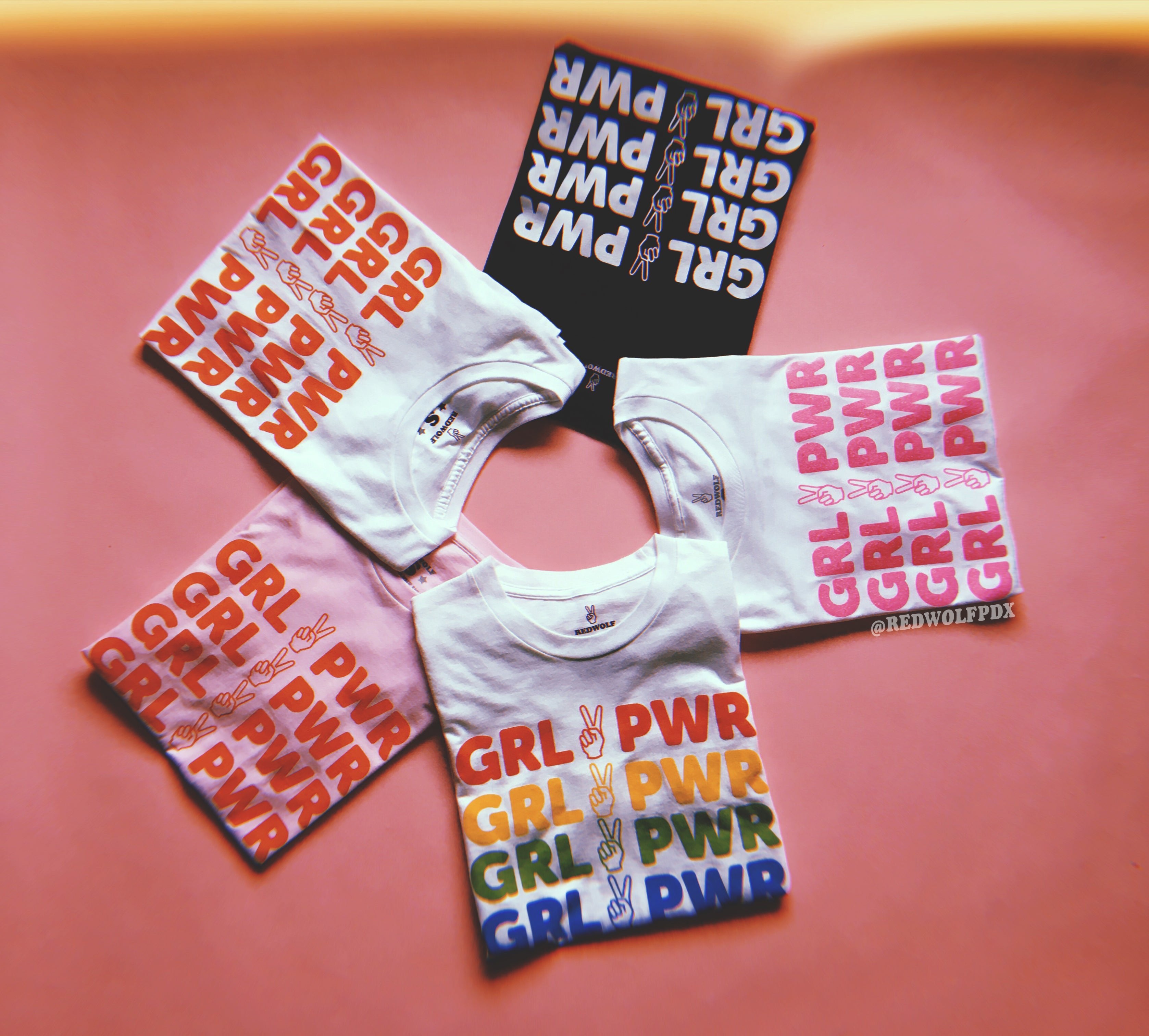 multiple tees with text 'GRL PWR' with peace sign graphic screen printed down the front on a pink background