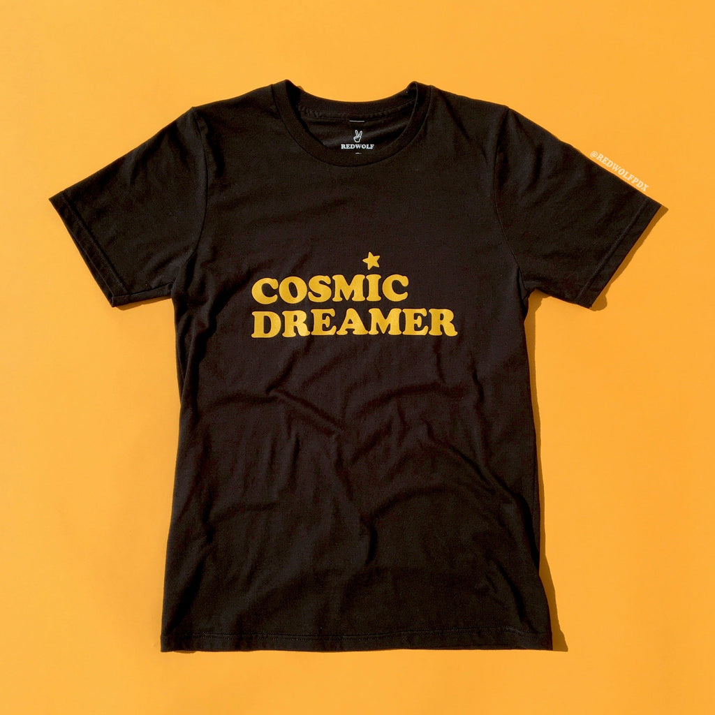 black graphic tee with text 'COSMIC DREAMER' in yellow ink screen printed across chest against a yellow backdrop