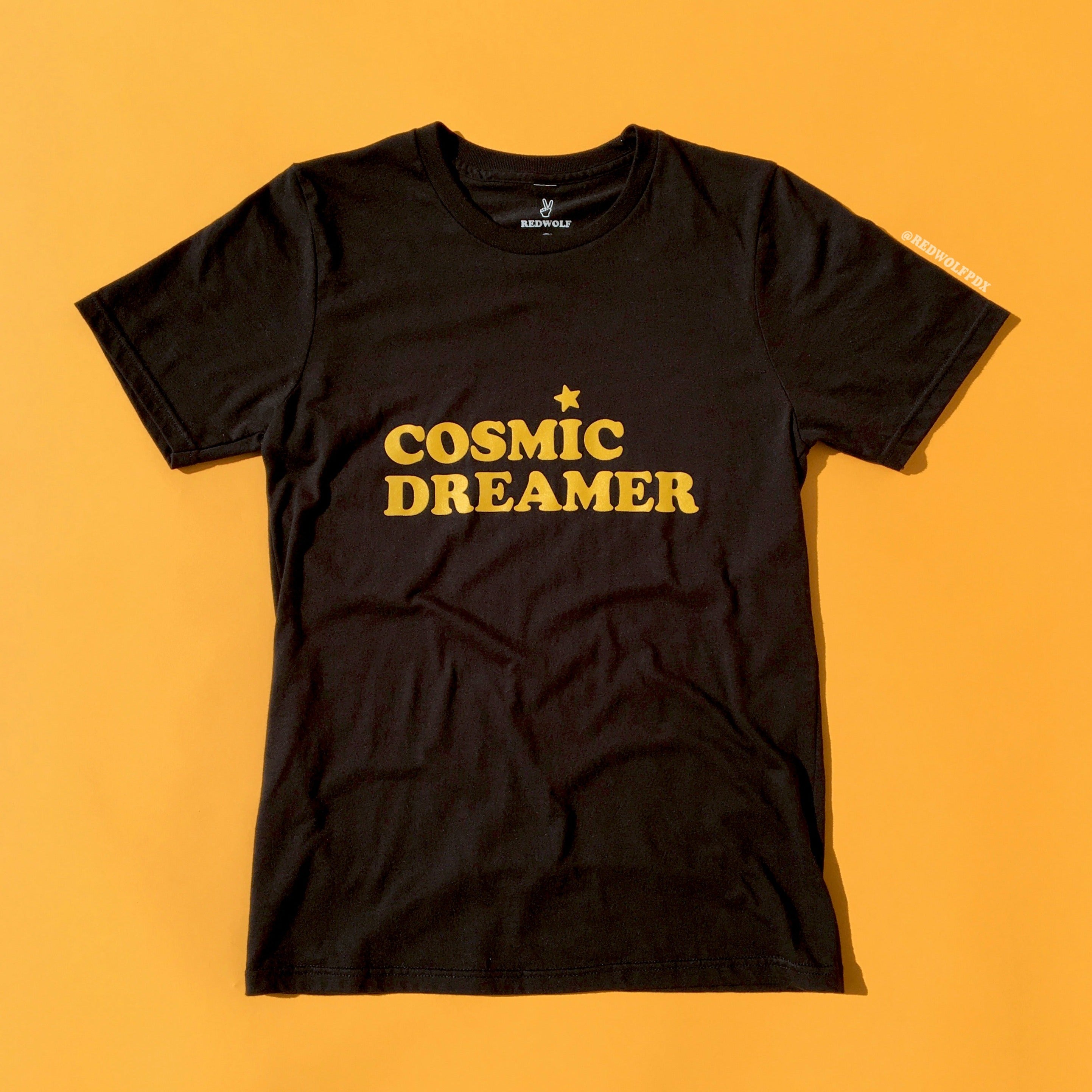 black graphic tee with text 'COSMIC DREAMER' in yellow ink screen printed across chest against a yellow backdrop