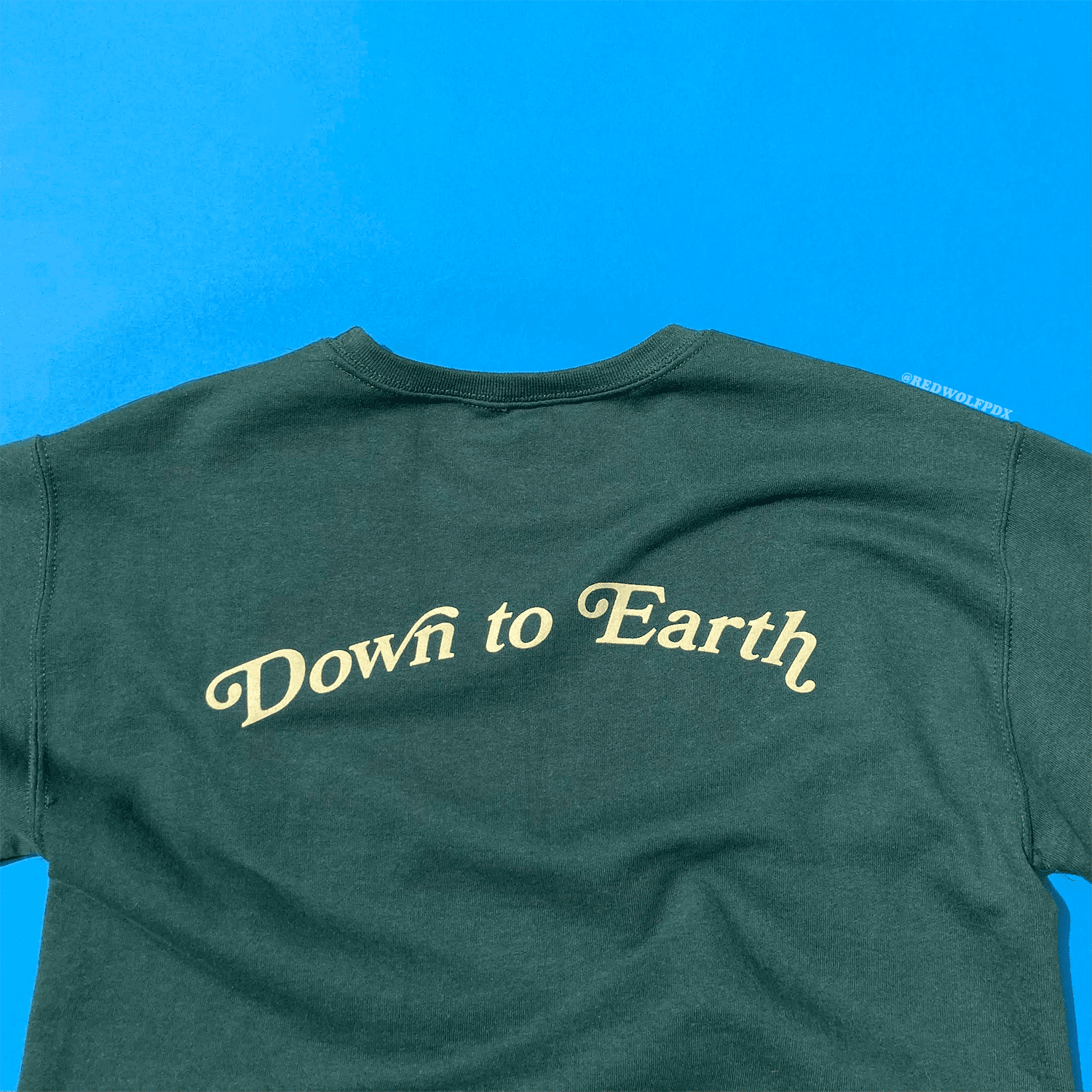 'DOWN TO EARTH' text graphic over forest green crewneck sweatshirt against blue backdrop