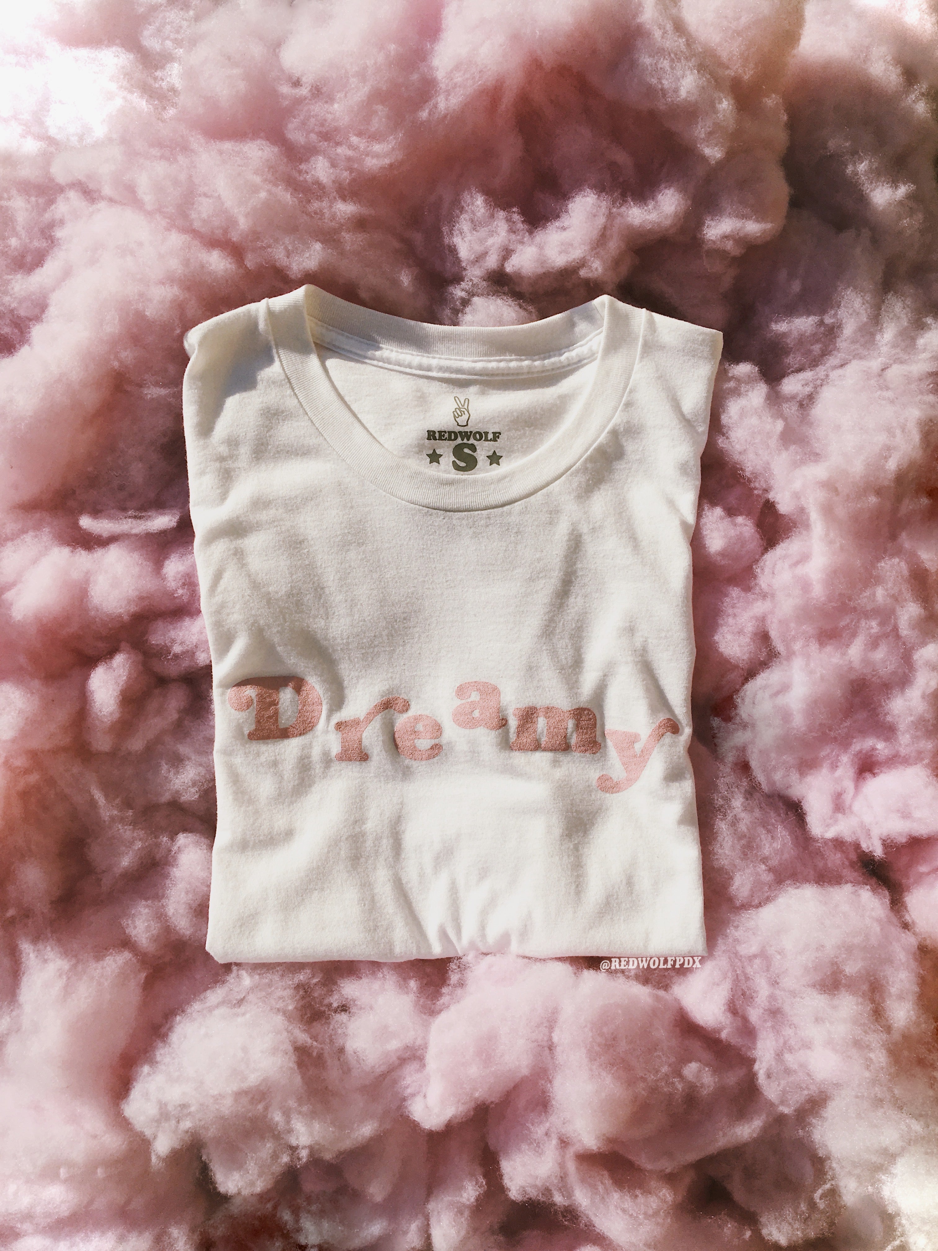 Folded white graphic tee with text 'Dreamy' in baby pink ink screen printed on front laying on fluffy pink cloud background