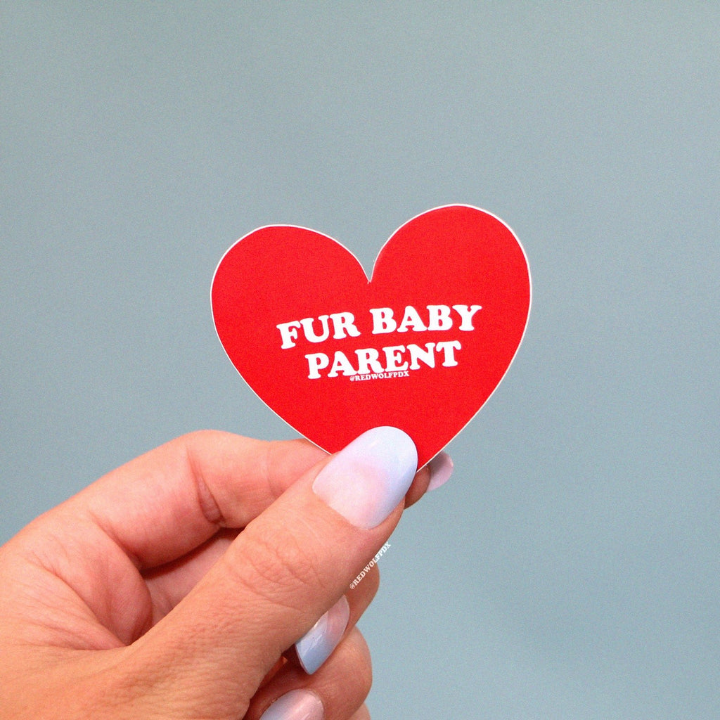 red heart shaped sticker with text 'FUR BABY PARENT' being held by hand against baby blue backdrop