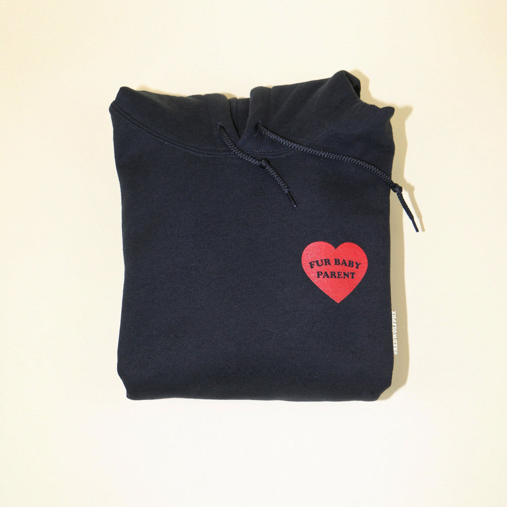 Folded black hoodie with illustrated heart with text 'FUR BABY PARENT' in red ink screen printed on front against a beige background