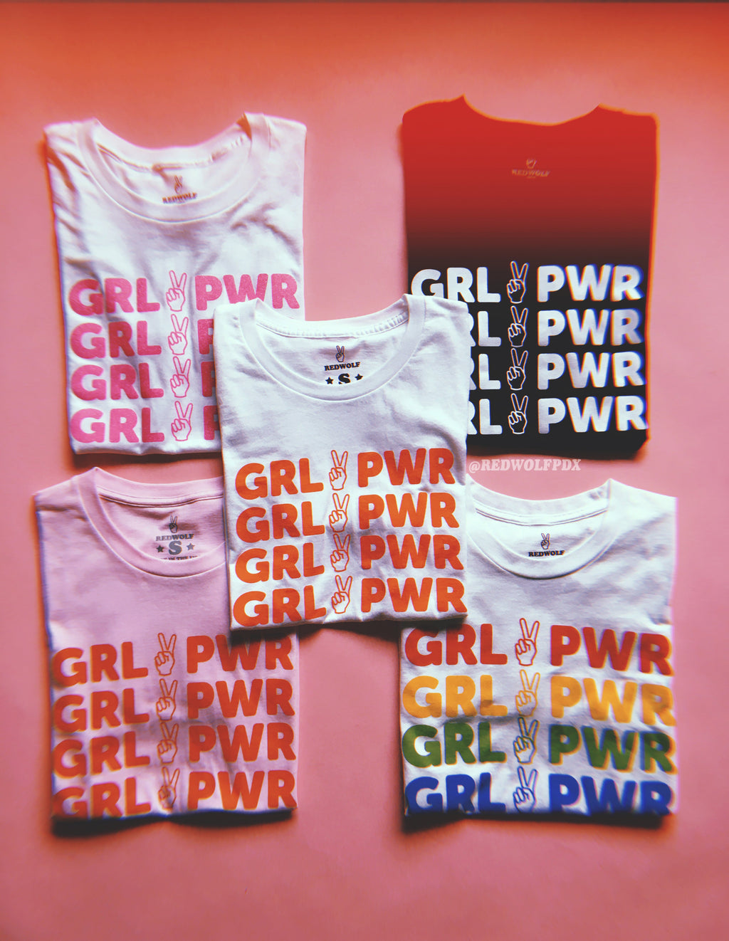 multiple folded tees with text 'GRL PWR' with peace sign graphic screen printed down the front on a pink background