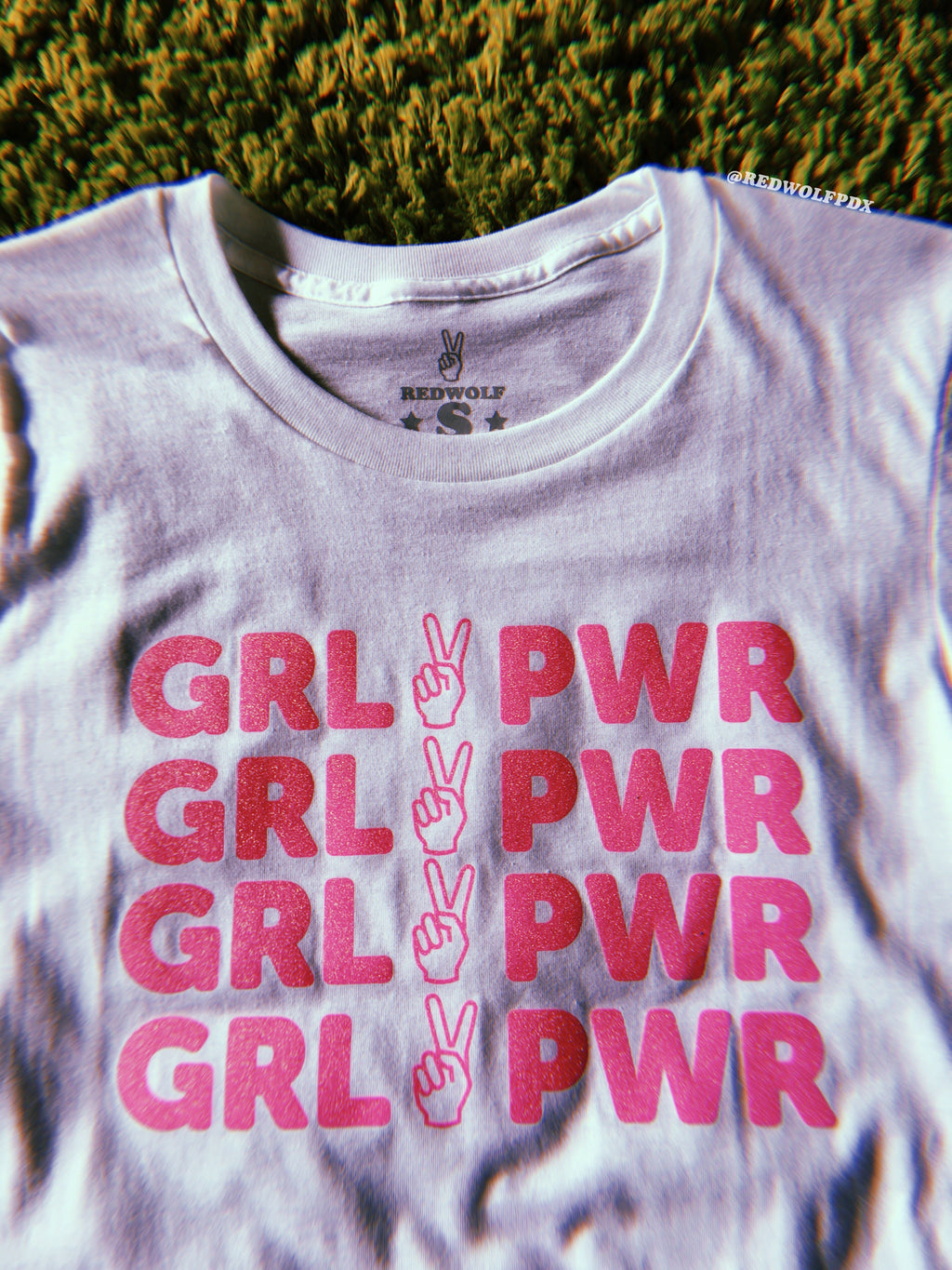 white tee with text 'GRL PWR' with peace sign graphic screen printed in pink glitter ink across the front against a vintage green background