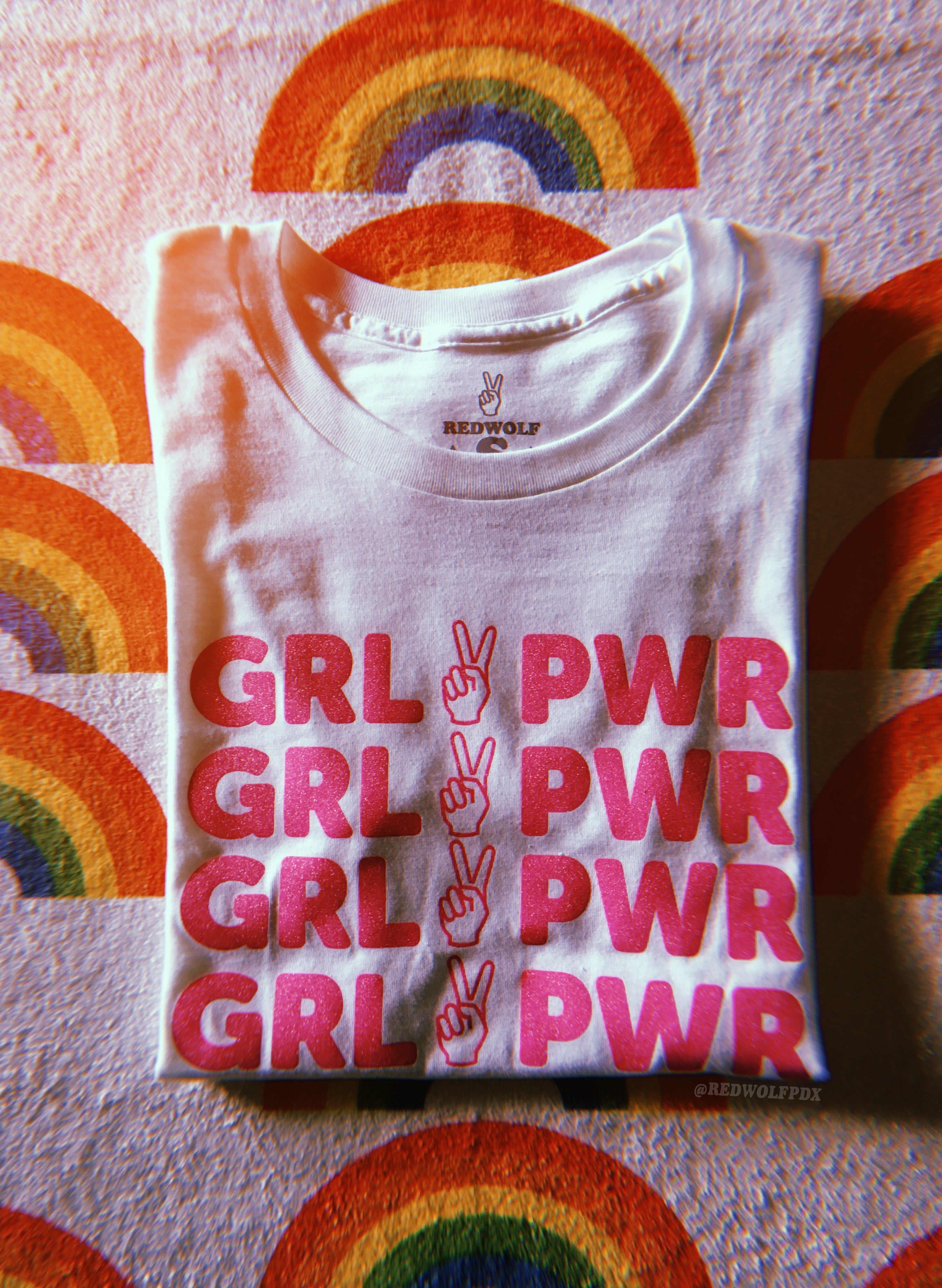 folded white tee with text 'GRL PWR' with peace sign graphic screen printed in pink glitter ink across the front on a rainbow print background
