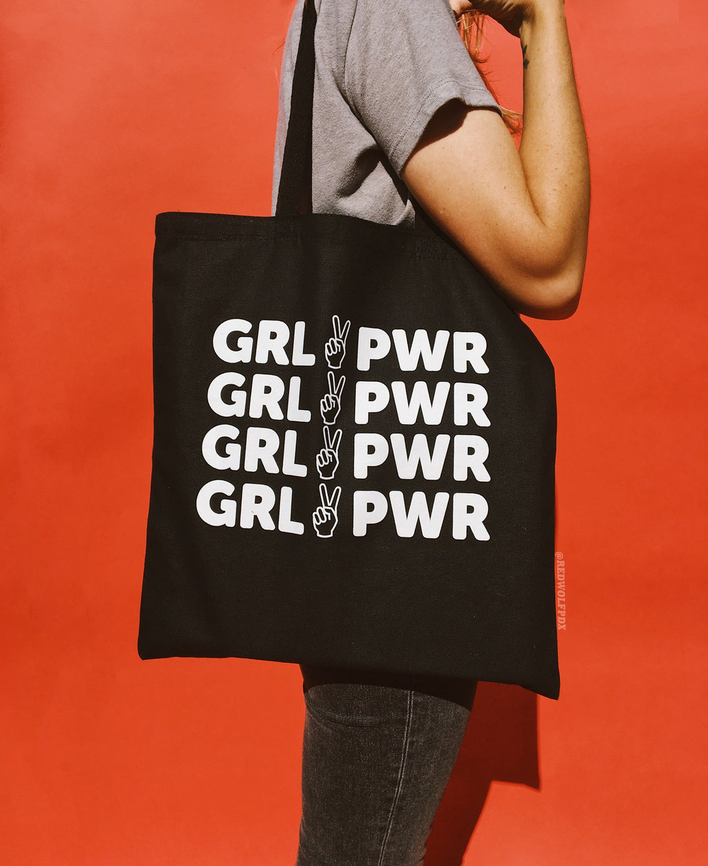 Black canvas tote bag with text 'GRL PWR' in white ink screen printed across front being worn on shoulder against a red backdrop