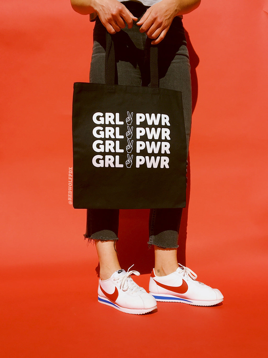 black canvas tote bag with text 'GRL PWR'  in white ink screen printed across tote being held against a red backdrop