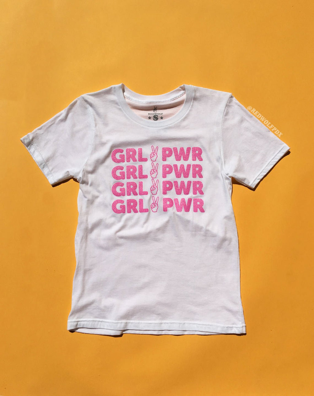 white tee with text 'GRL PWR' with peace sign graphic screen printed in pink glitter ink across the front against a  yellow background