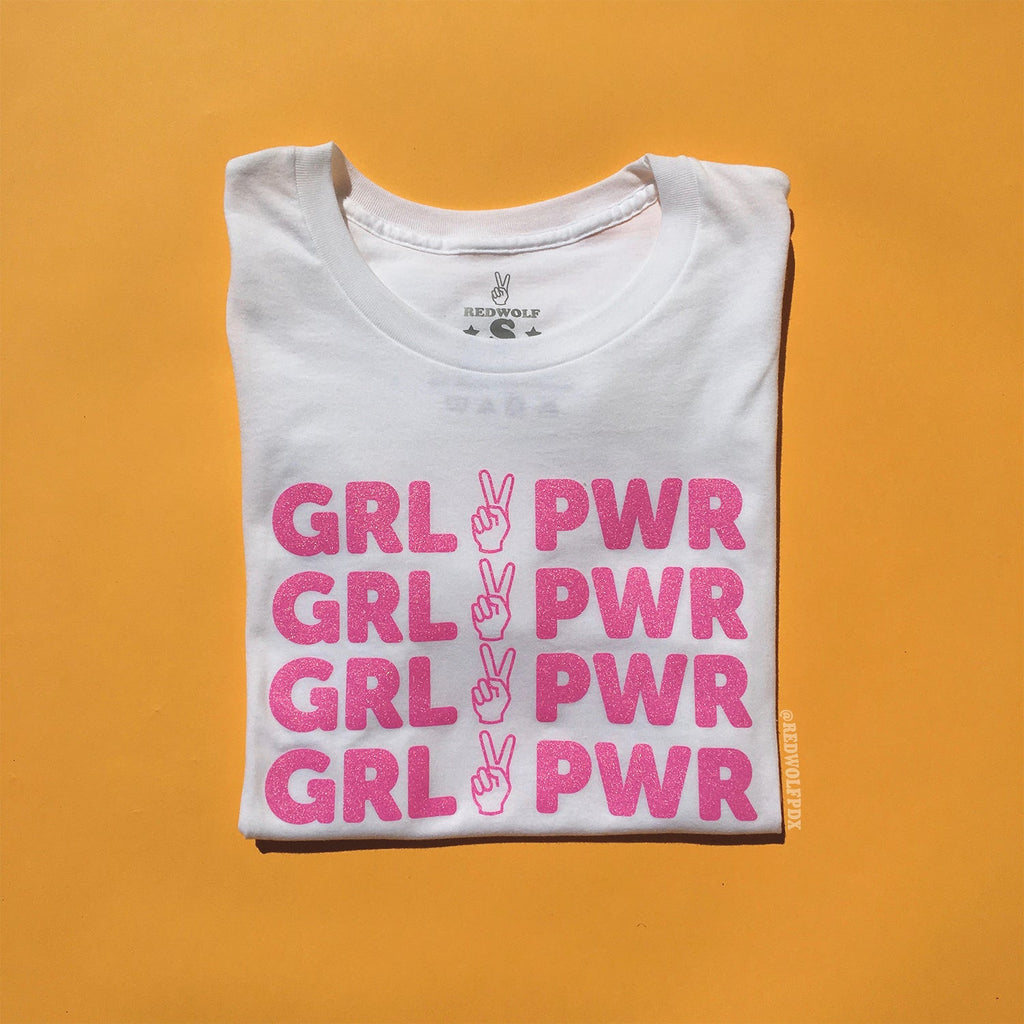 folded white tee with text 'GRL PWR' with peace sign graphic screen printed in pink glitter ink across the front on a yellow background