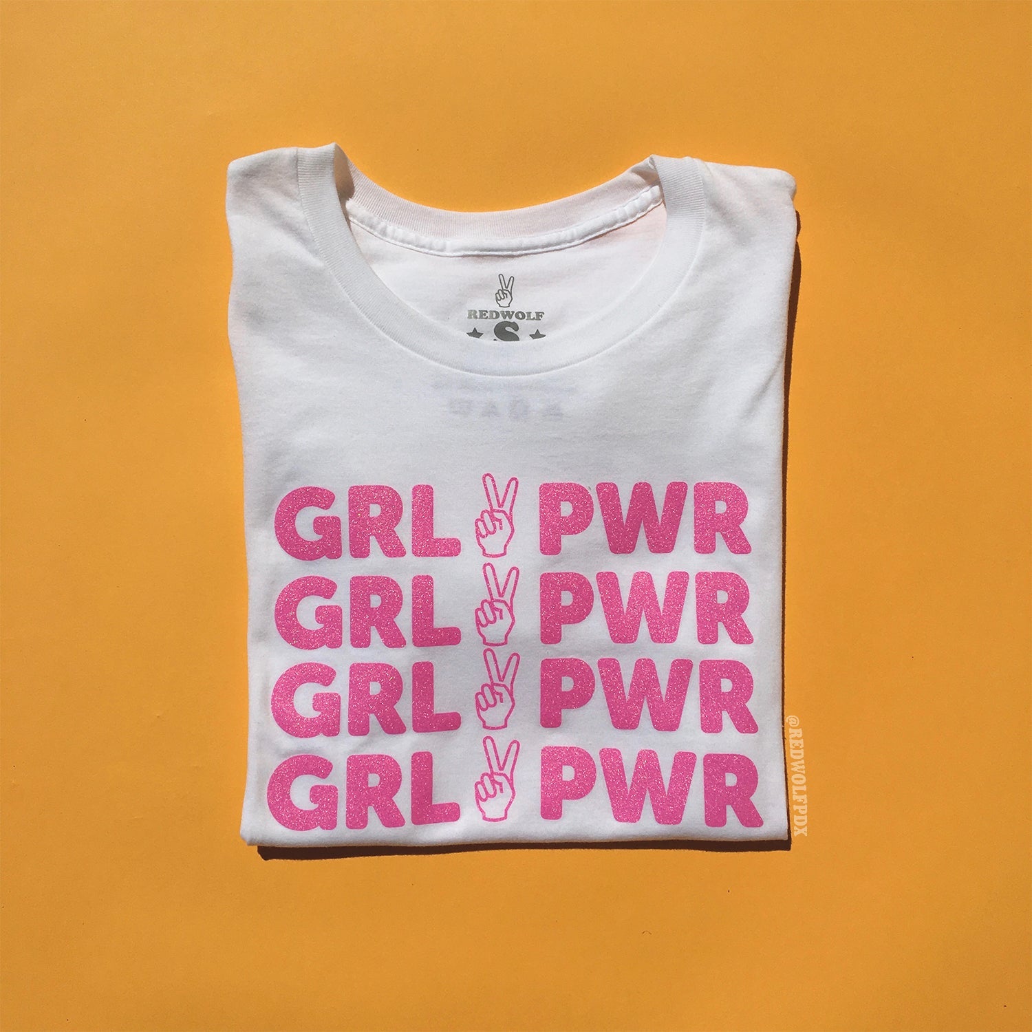 folded white tee with text 'GRL PWR' with peace sign graphic screen printed in pink glitter ink across the front on a yellow background