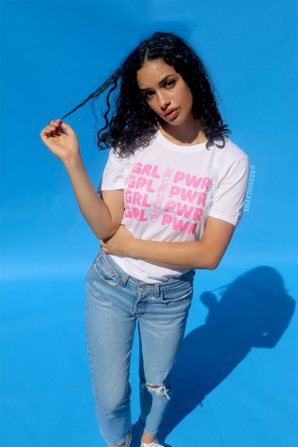 white tee with text 'GRL PWR' with peace sign graphic screen printed in pink glitter ink across the front being worn against a blue backdrop