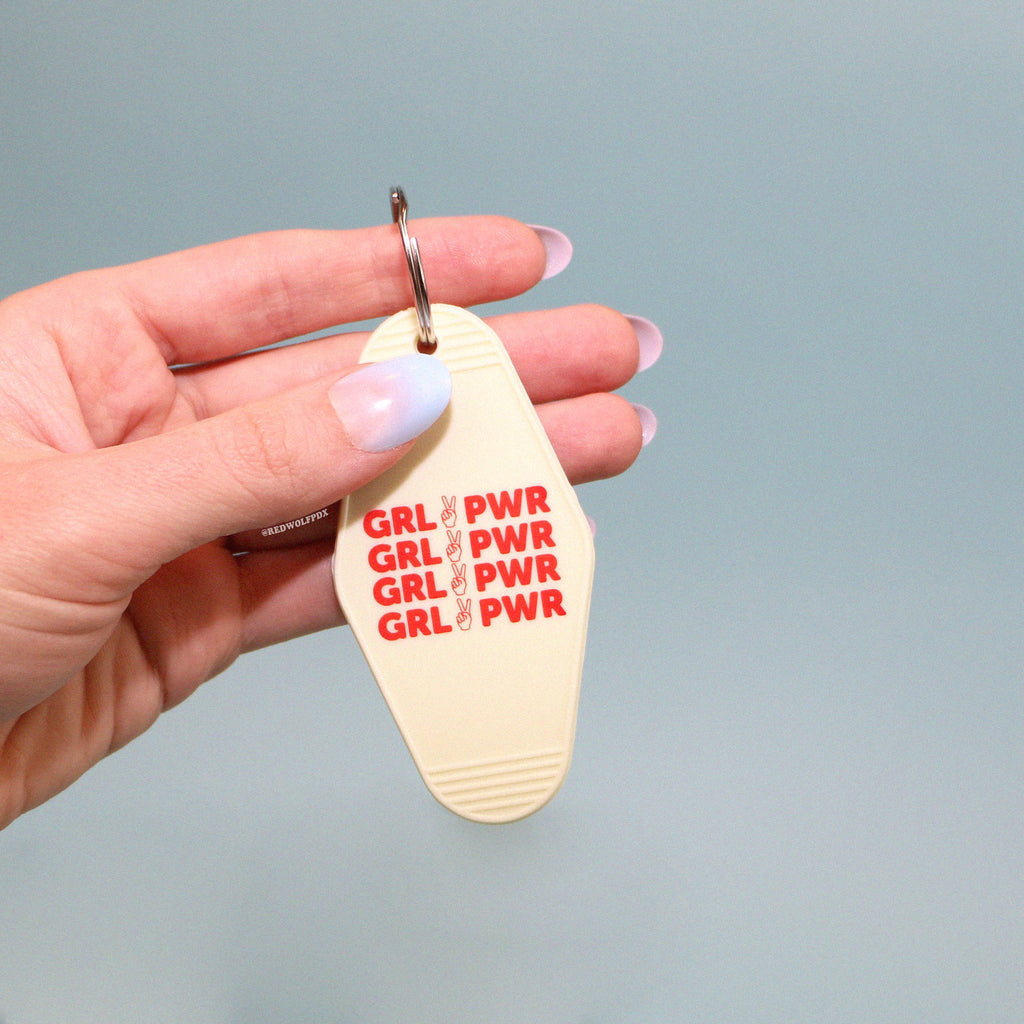 A vintage white keychain with the text 'GRL PWR' in red being held against a blue backdrop