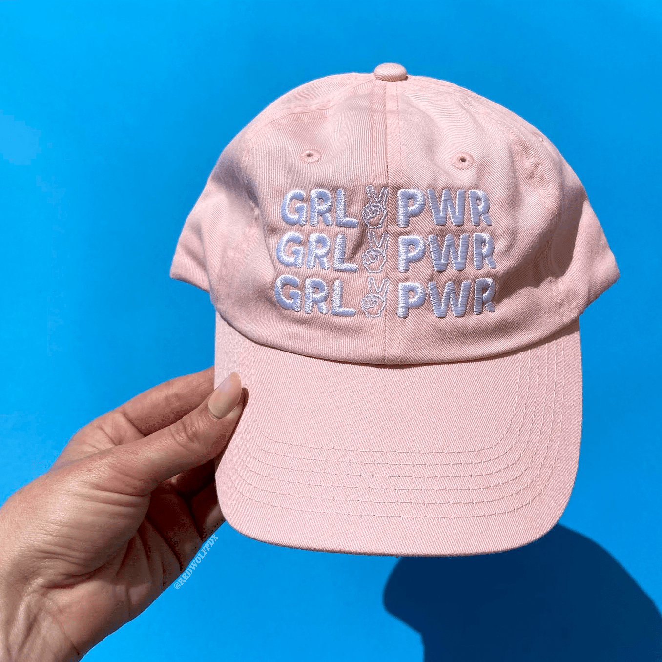 Baby pink baseball cap with text 'GRL PWR' embroidered in white thread across front being held against a blue backdrop