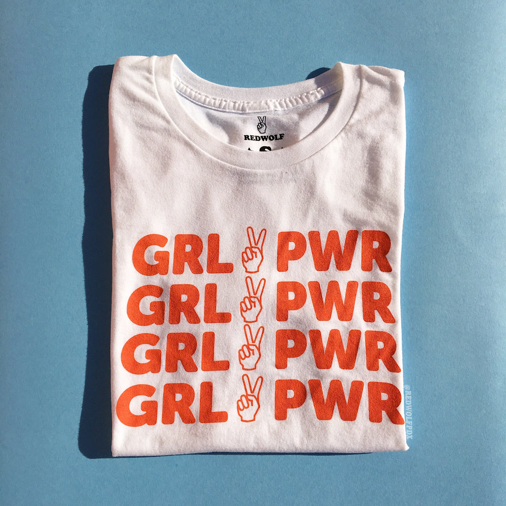 folded white tee with text 'GRL PWR' with peace sign graphic in vintage red orange ink screen printed down the front on a light blue background