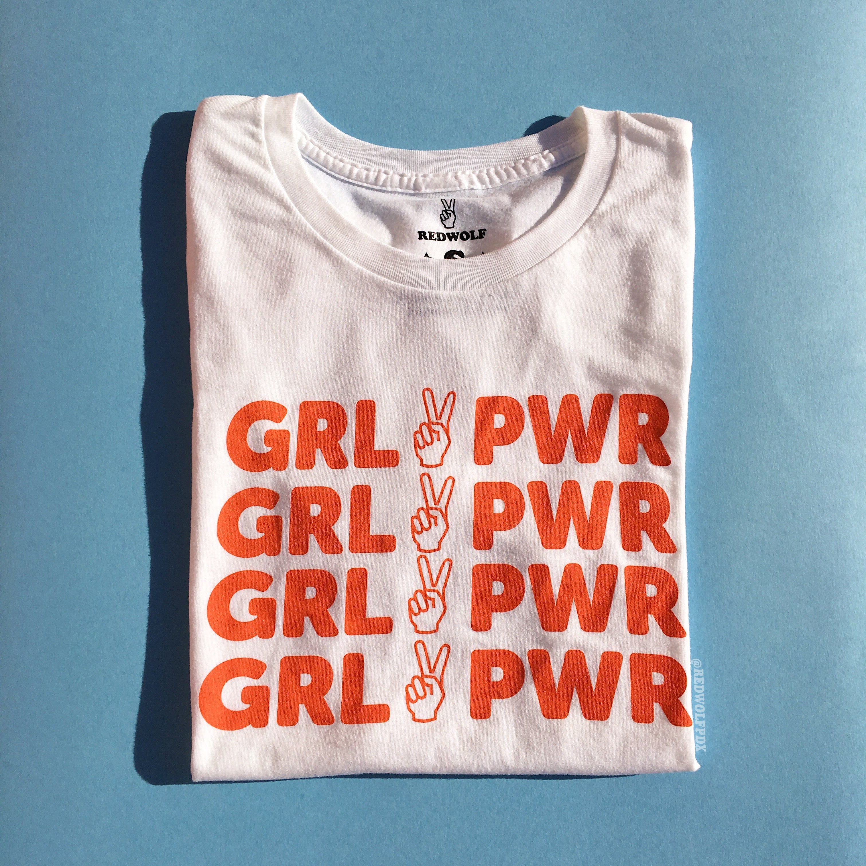 folded white tee with text 'GRL PWR' with peace sign graphic in vintage red orange ink screen printed down the front on a light blue background