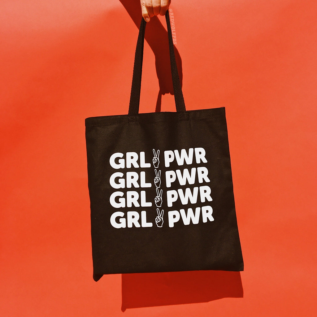 Black canvas tote bag with text 'GRL PWR' in white ink screen printed across being held up against a vintage red backdrop