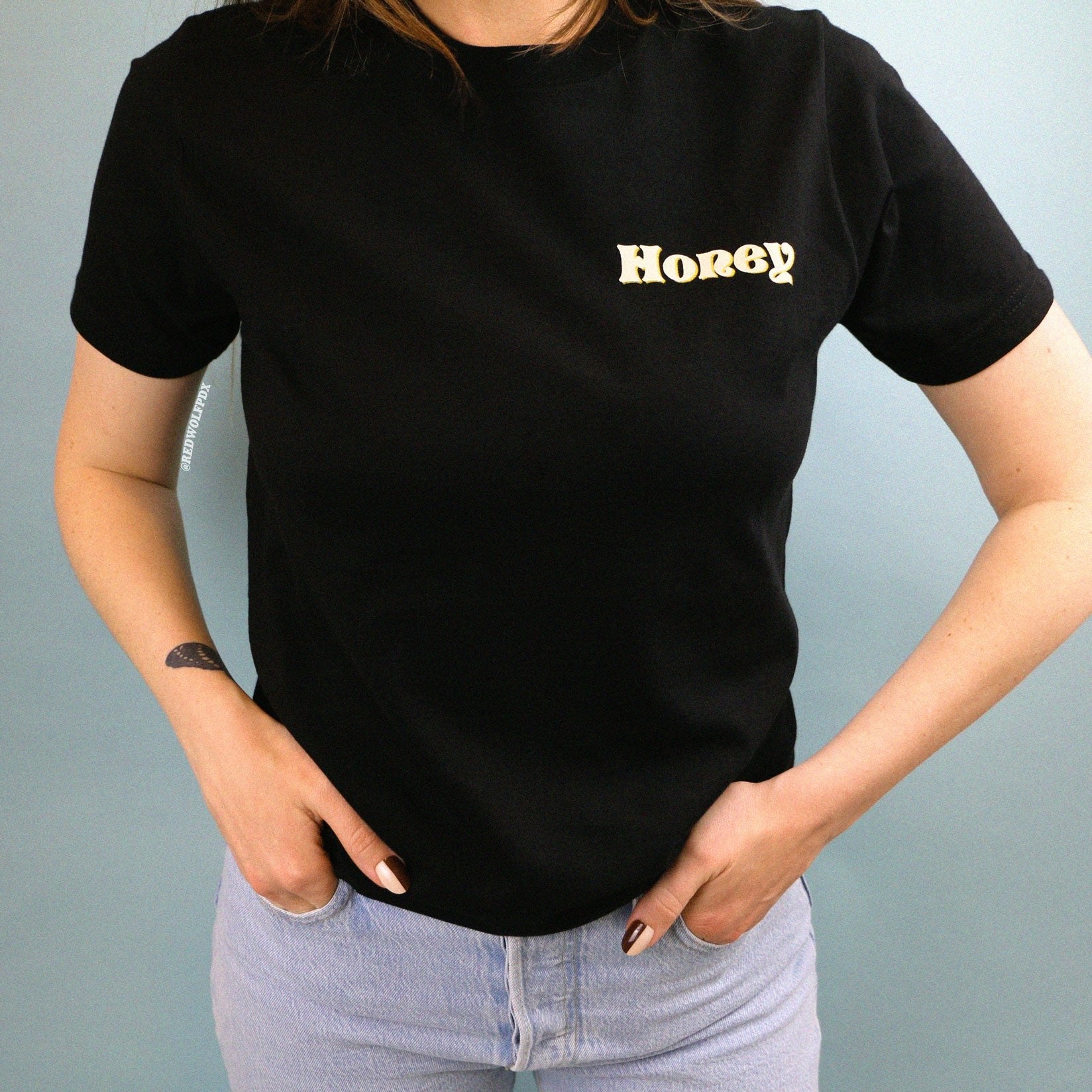 Black graphic tee with text 'Honey' with layered white and yellow ink screen printed on front corner chest being worn against a blue backdrop.