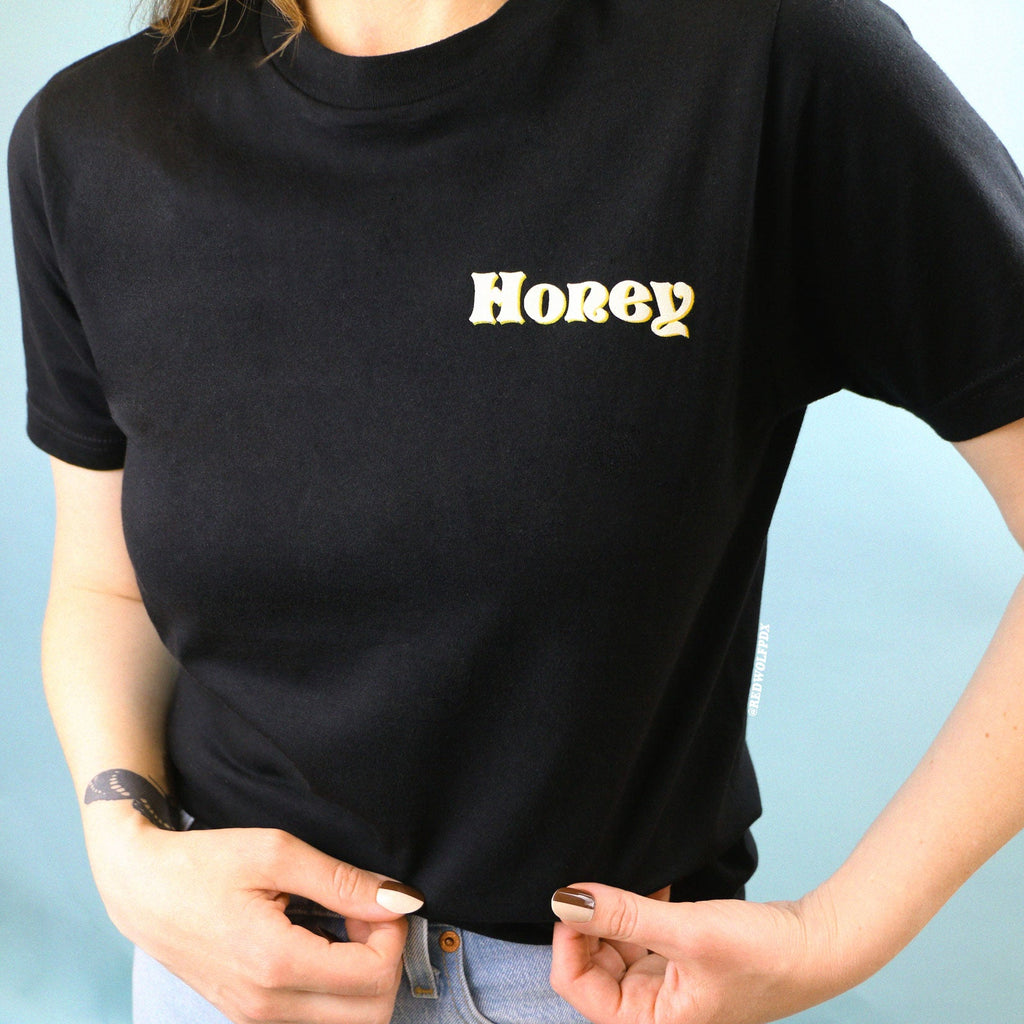 Black graphic tee with text 'Honey' in white ink shadowed with yellow ink screen printed on front corner of chest being worn against blue backdrop
