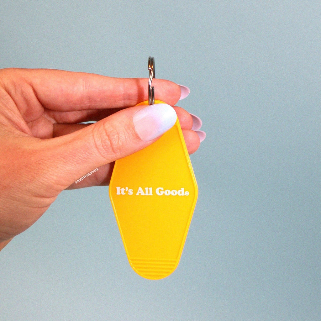 yellow gold vintage motel style keychain with text 'It's All Good' with smiley face illustration graphic across front being held against a light blue backdrop