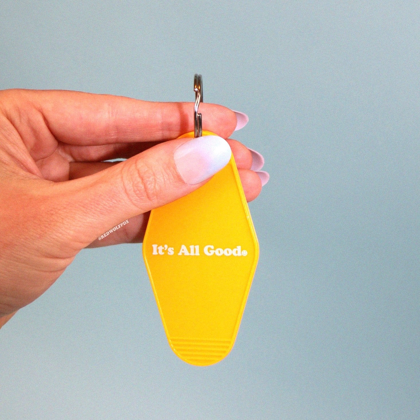 yellow gold vintage motel style keychain with text 'It's All Good' with smiley face illustration graphic across front being held against a light blue backdrop